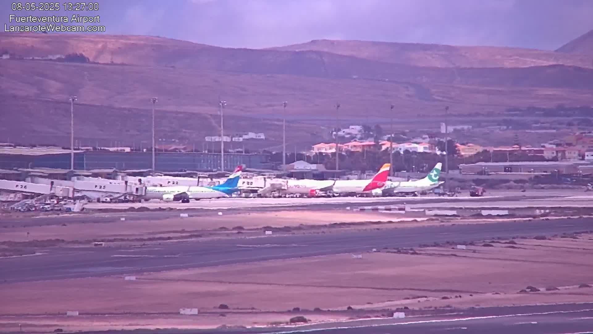 Several airplanes are parked at an airport tarmac under an overcast sky, with distant brown hills in the background.