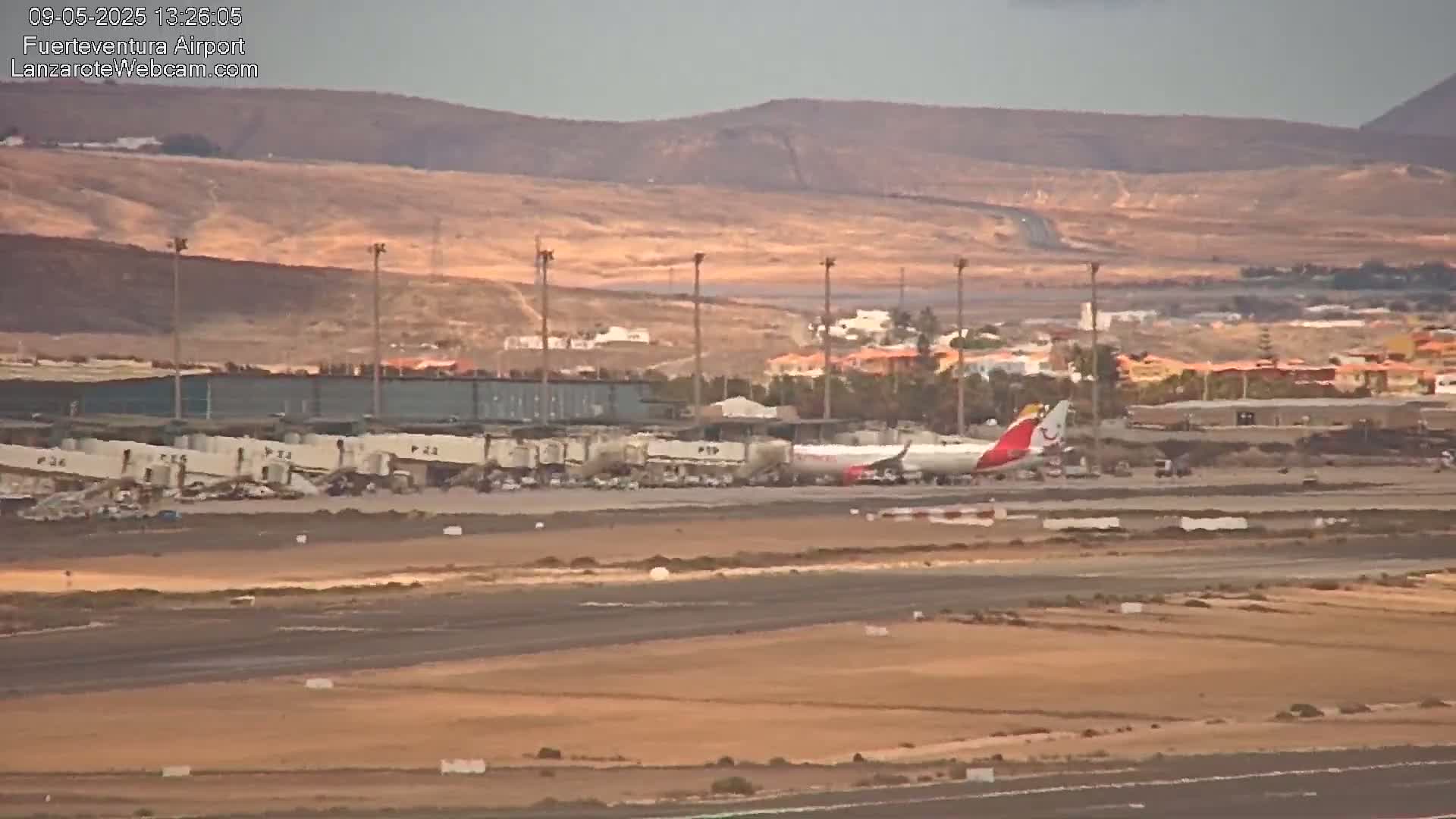 A plane is parked at an airport tarmac, near a terminal building, against a backdrop of brown hills under a mostly clear sky.