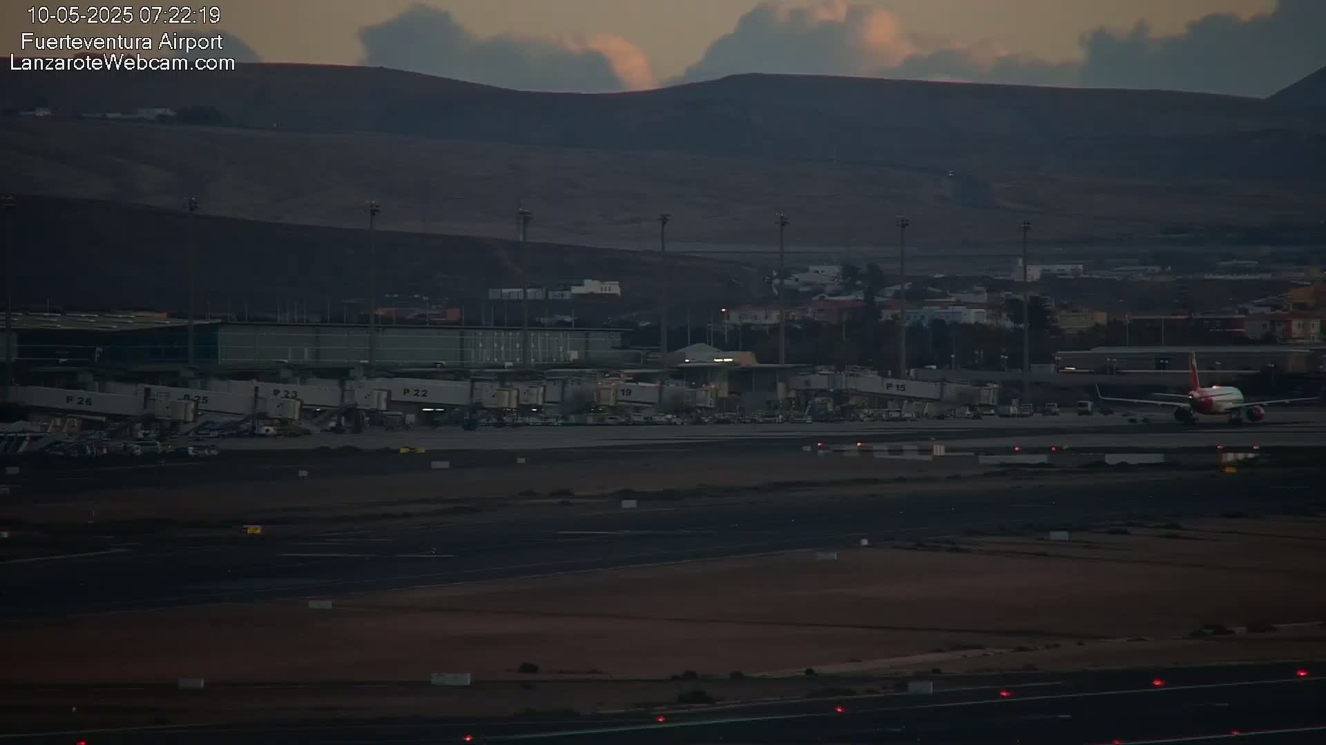 An airplane sits on a tarmac at an airport under a dusky, cloudy sky near some low mountains.