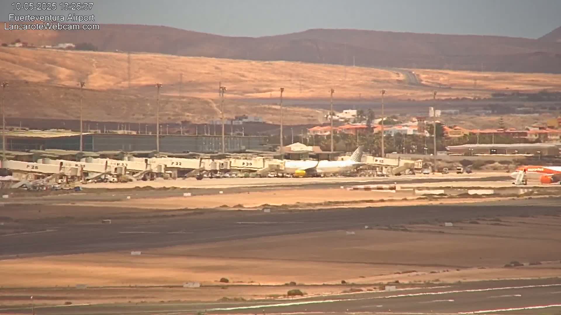 A wide shot of an airport tarmac under sunny conditions shows several airplanes parked at gates, with a distant view of brown hills and sparse buildings.