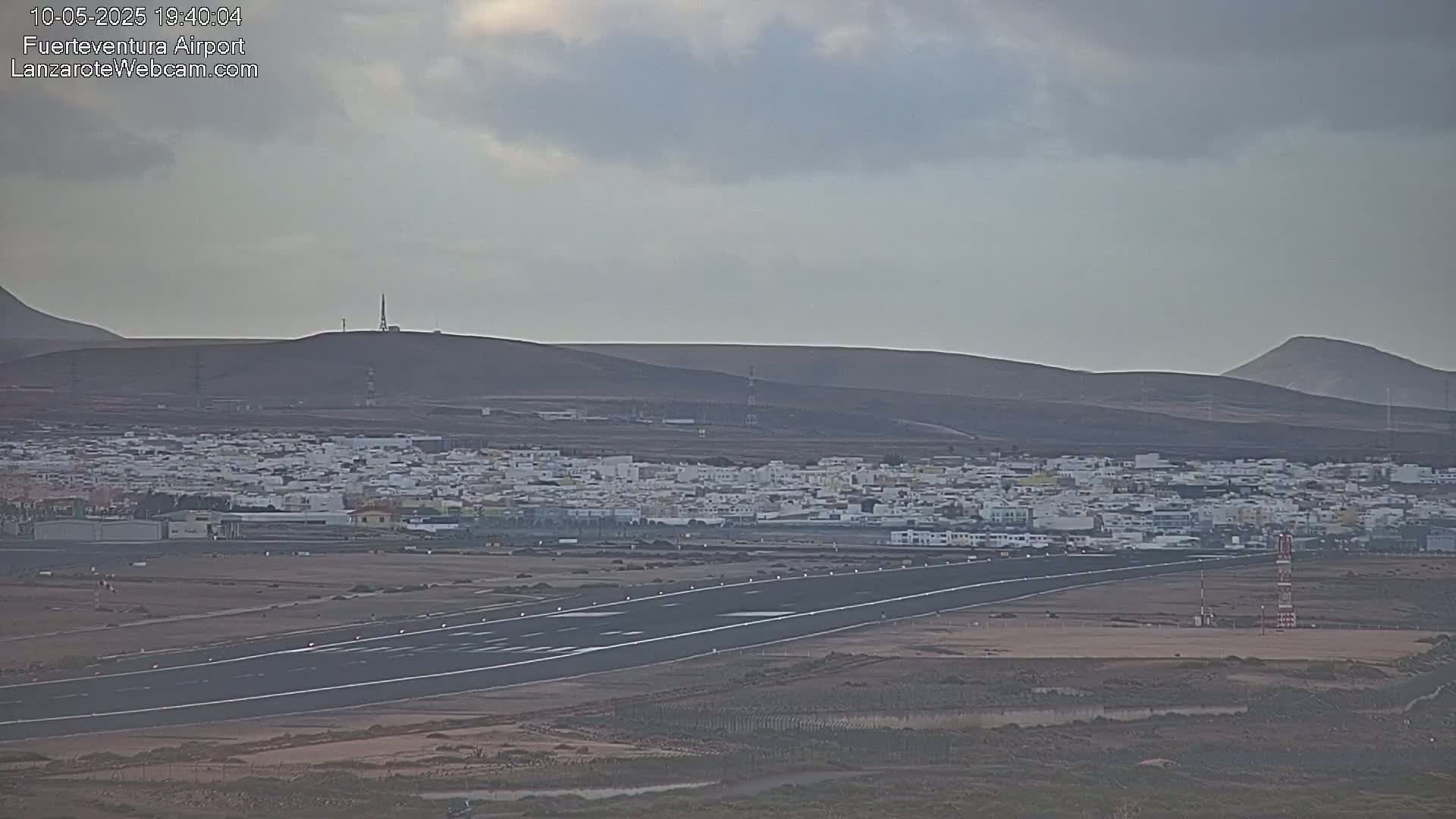 An airport runway stretches toward a town nestled between arid, brown hills under a cloudy sky.