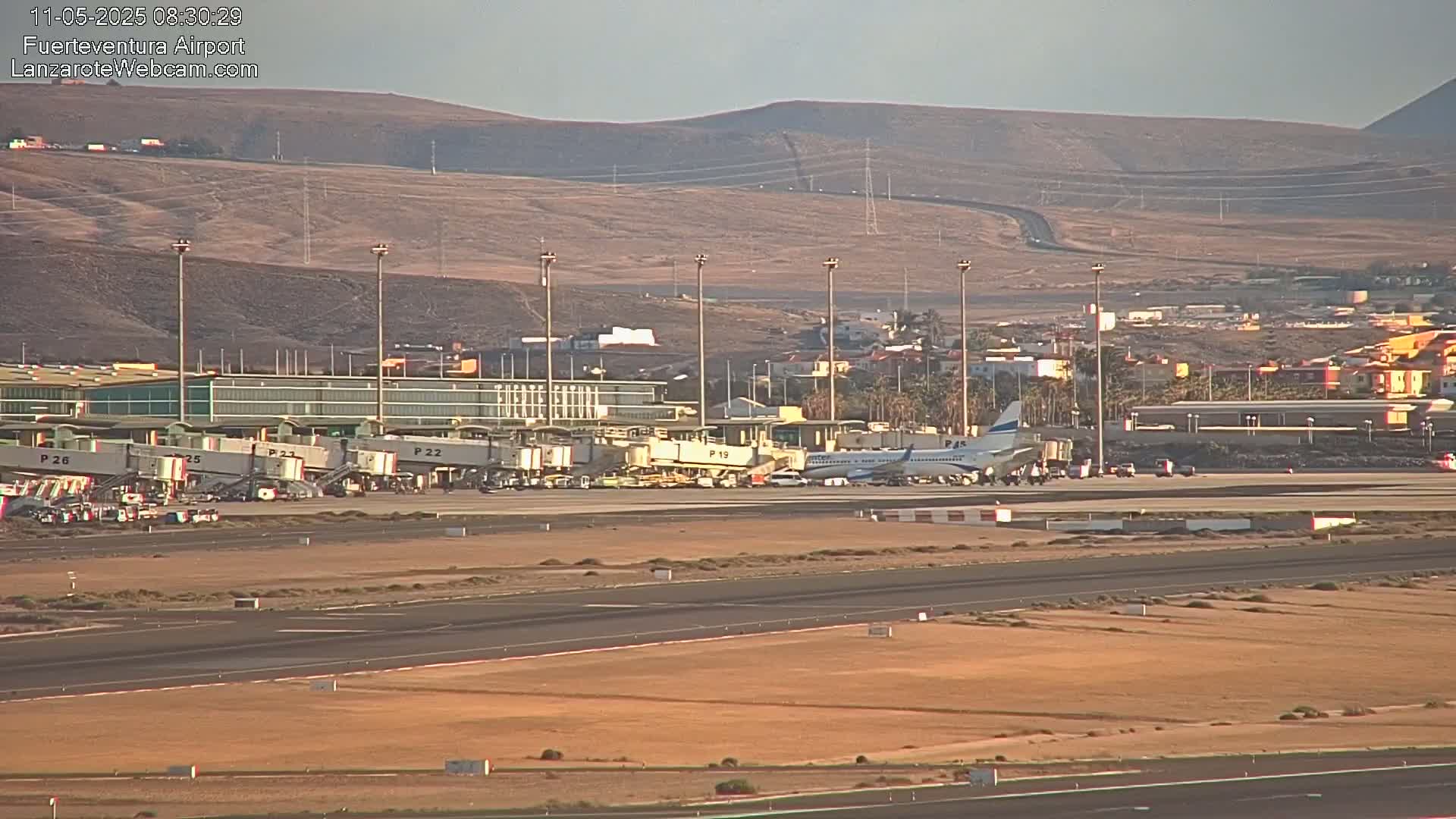 An airplane is parked at an airport terminal under a clear sky, with arid hills visible in the background.