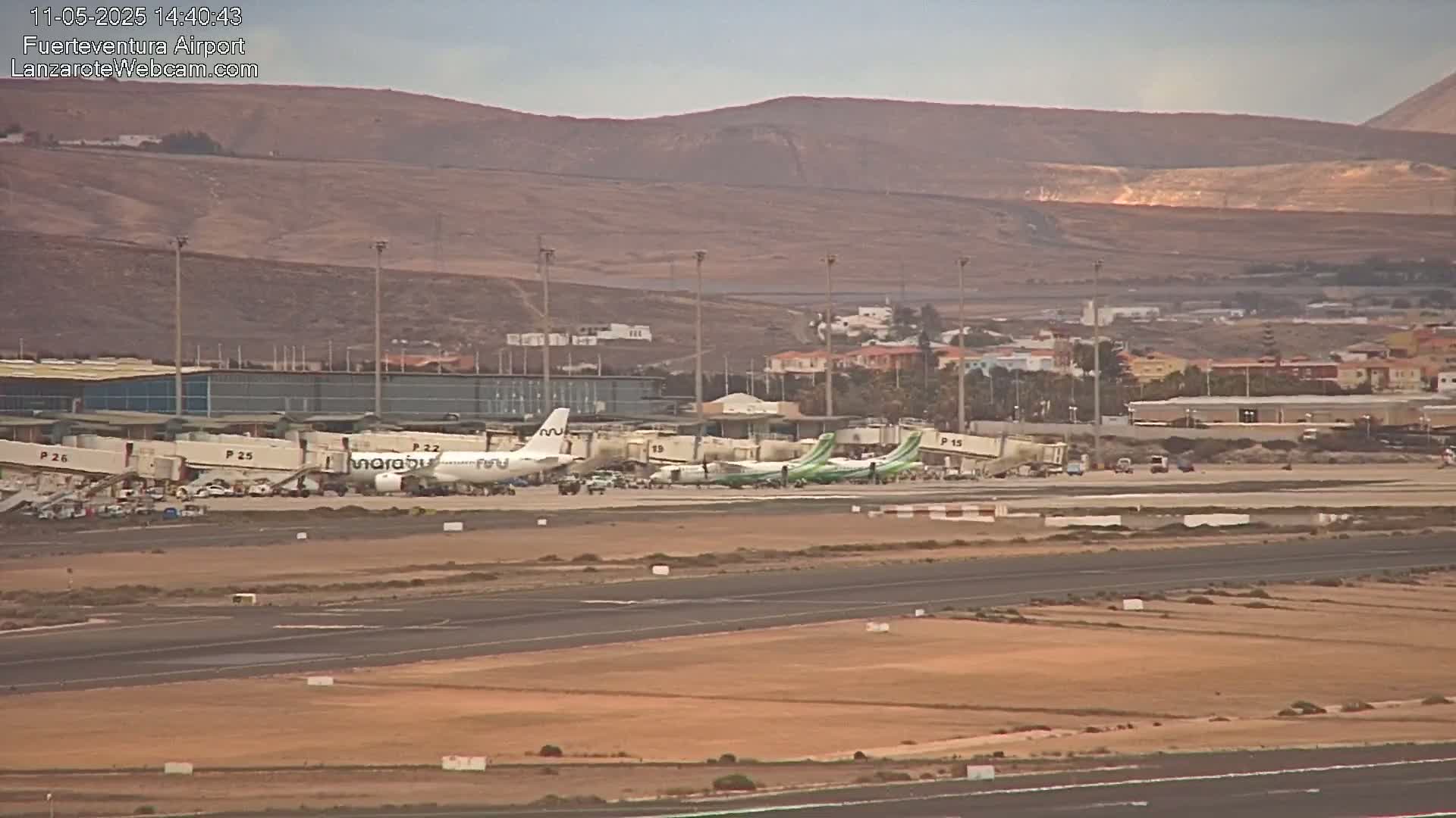 Several airplanes are parked at gates at an airport under a mostly clear sky, with brown hills visible in the distance.