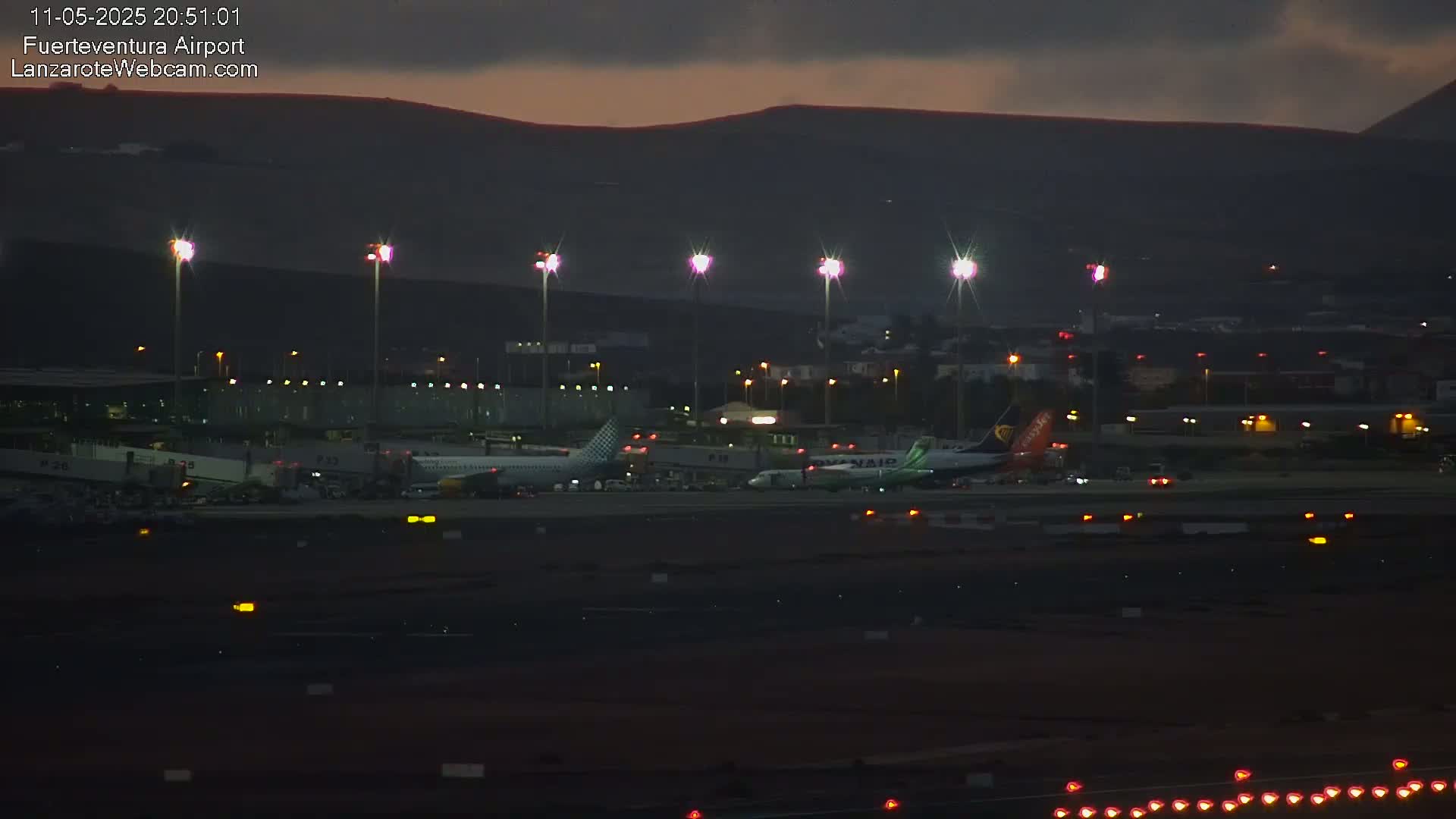 At twilight, several airplanes are parked on a dark airport tarmac, illuminated by bright lights against a backdrop of dark hills.