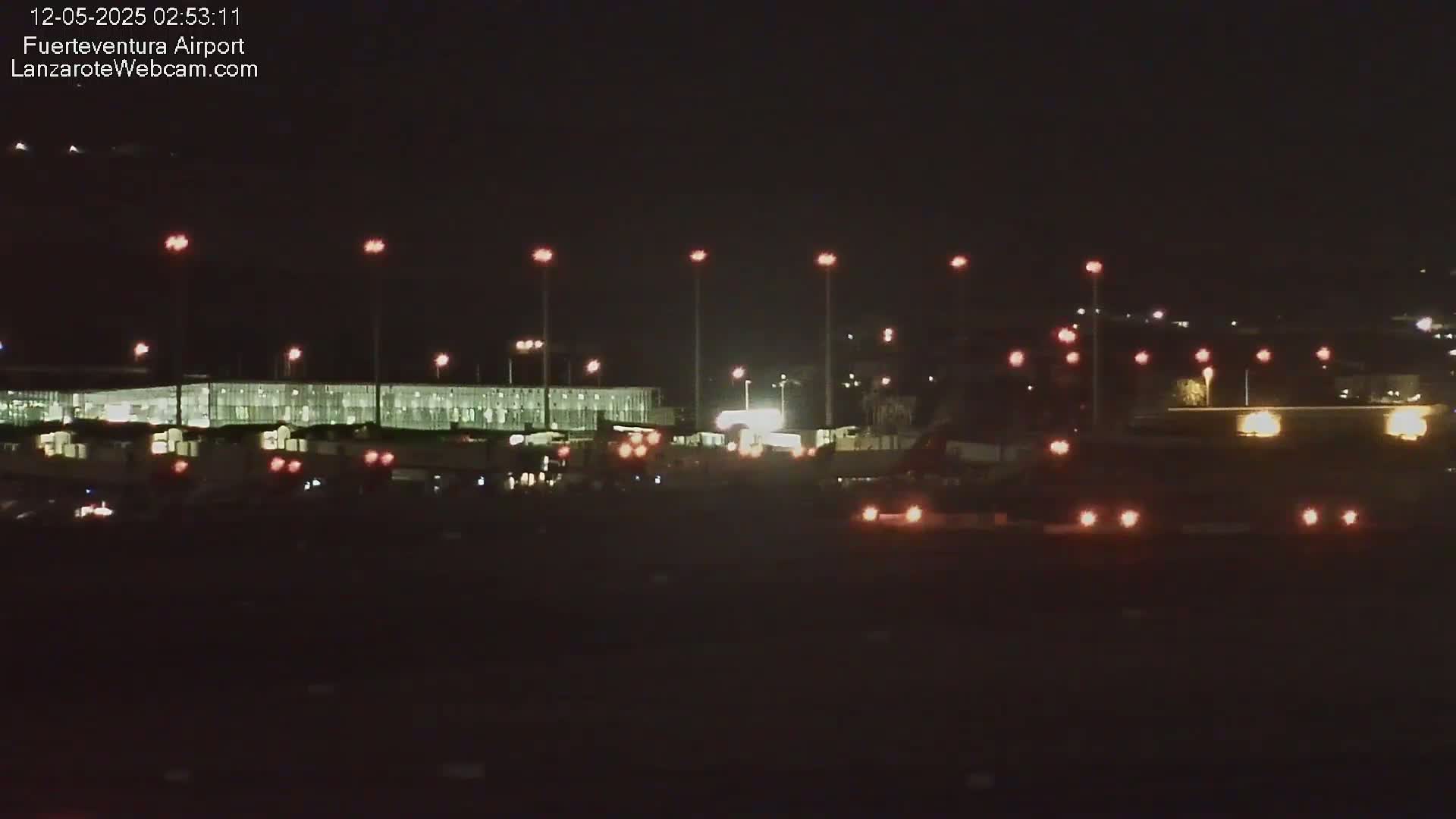 A nighttime view of an airport terminal building illuminated by lights, with vehicles visible on the tarmac under a dark sky.