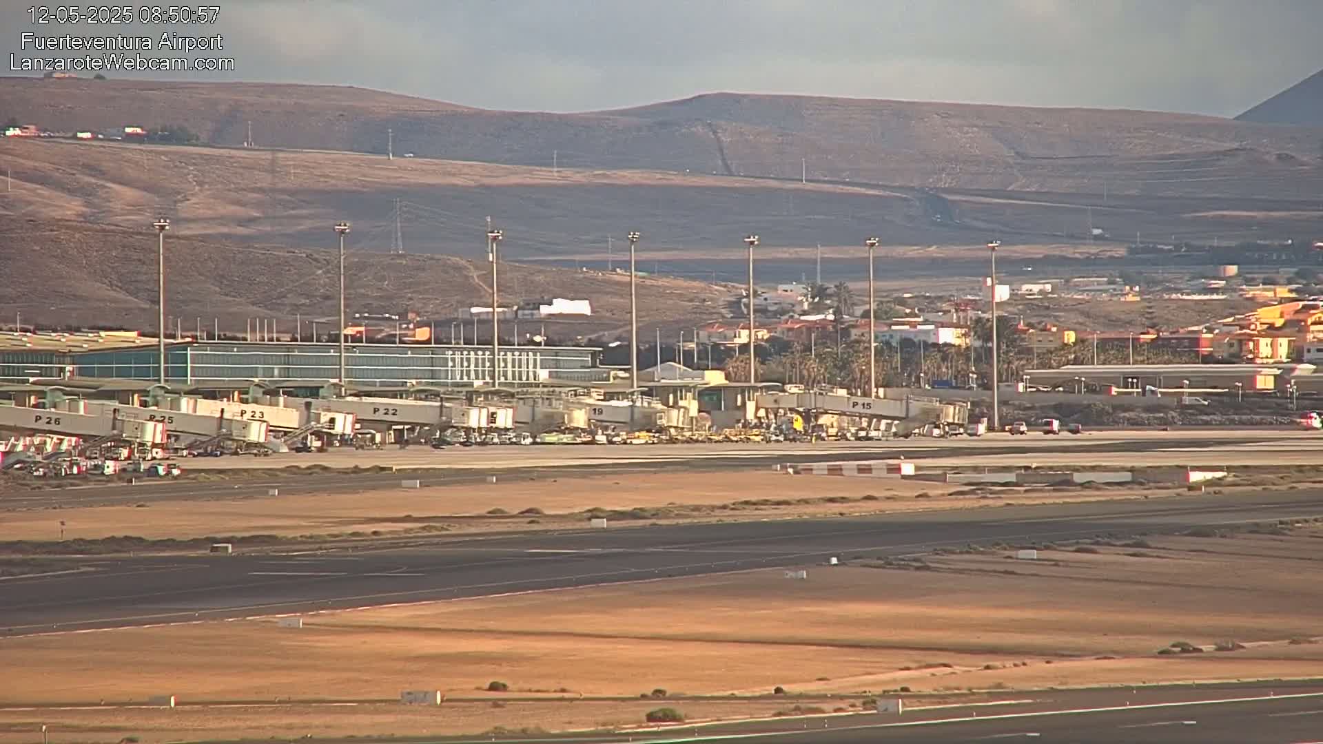 An airport terminal with several passenger bridges connected to airplanes is visible, under a mostly clear sky, with hills in the background.