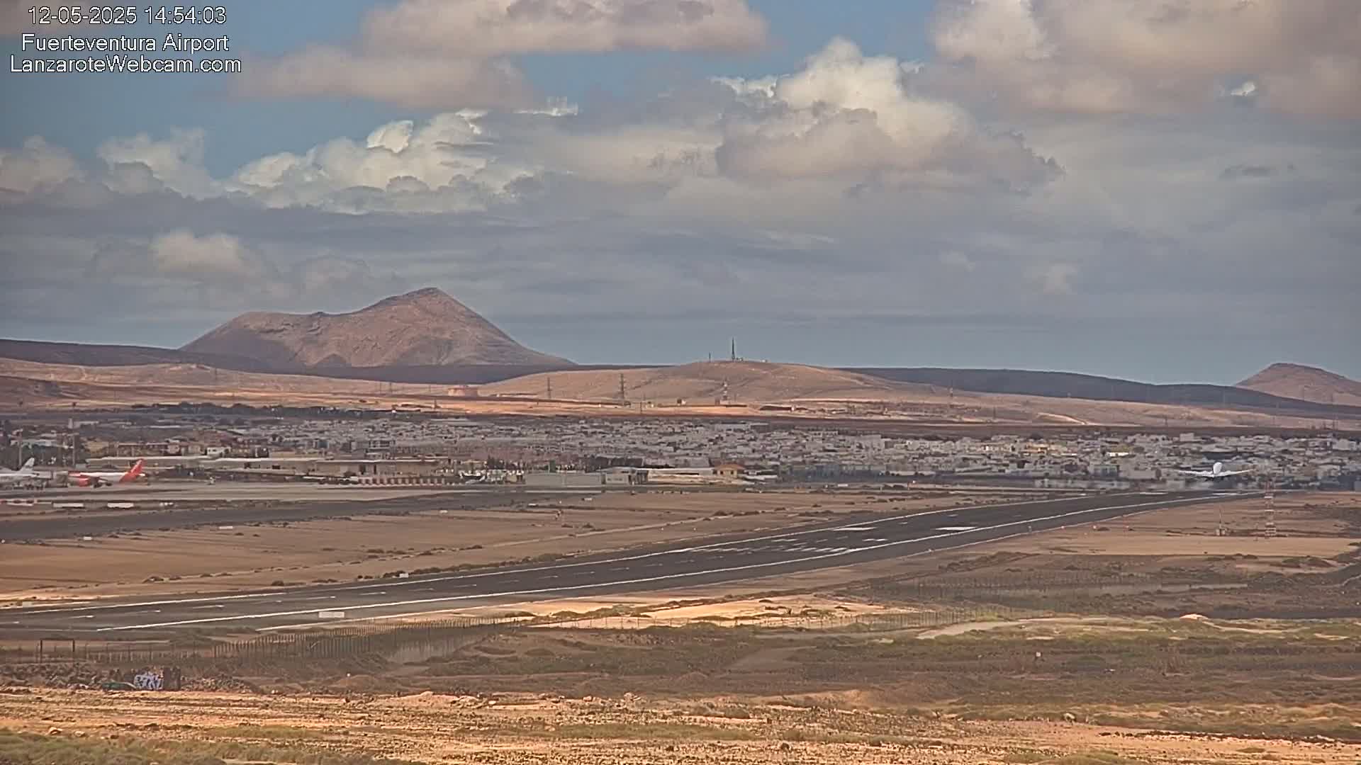 A long runway at an airport is visible, with planes on the tarmac, a town in the mid-ground, and several hills under a partly cloudy sky.