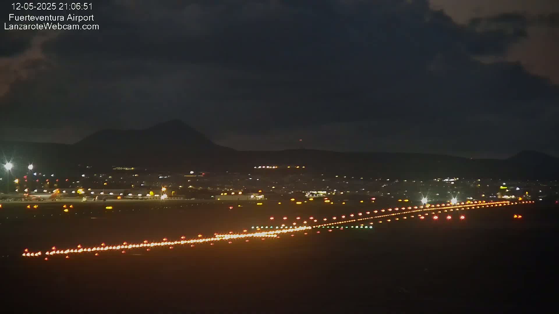 A nighttime view of an airport runway with lights illuminating the tarmac, under a dark, cloudy sky with hills in the background.