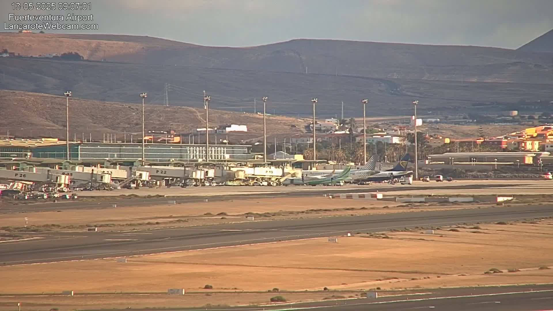 An airplane is parked at an airport tarmac with several jet bridges connected to a terminal building under a mostly clear sky, with brown, hilly terrain visible in the background.
