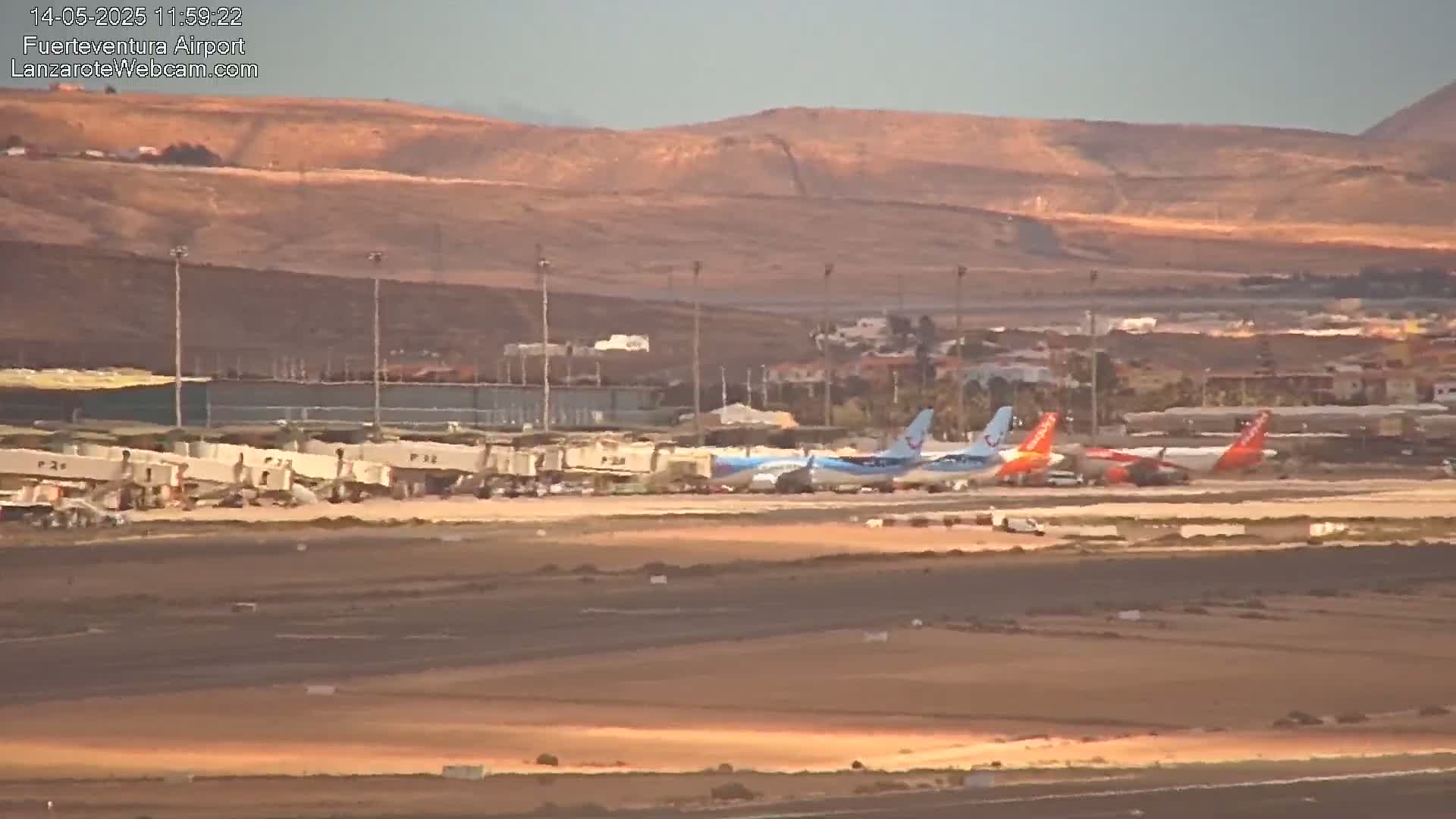 Several airplanes are parked at an airport tarmac under a clear, sunny sky, with brown hills visible in the background.