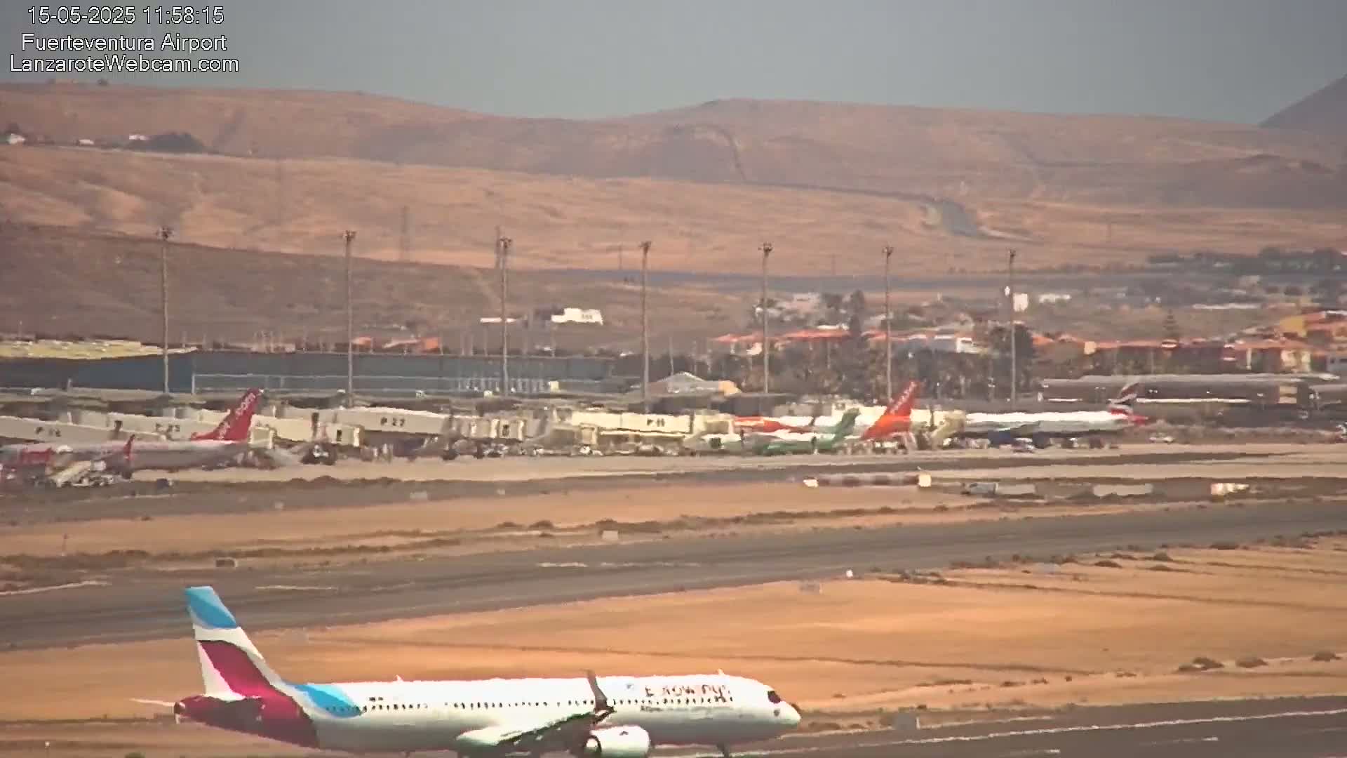 A plane taxis on an airport runway, with several other planes parked nearby, under a hazy sky, against a backdrop of arid hills.