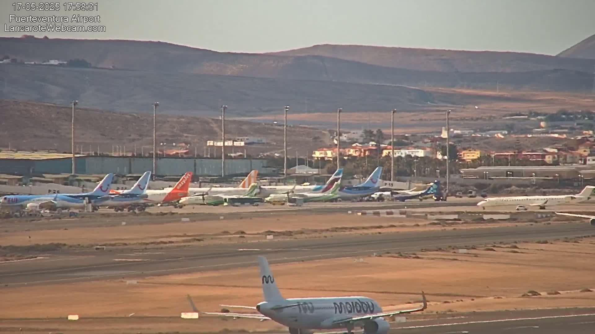 Many airplanes are parked on a tarmac at an airport under a hazy sky, with a single plane taxiing on the runway in the foreground.