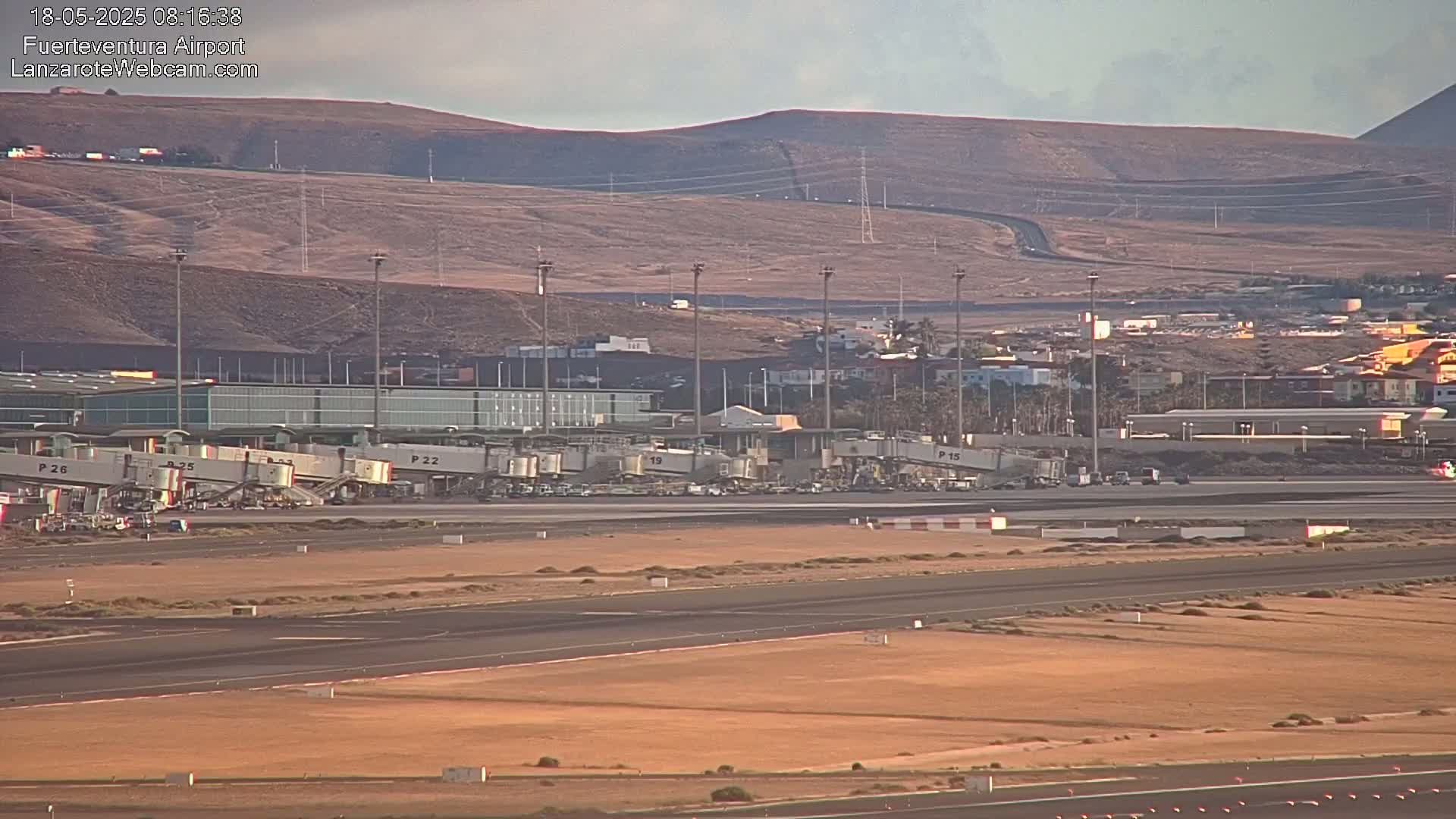 An airport tarmac with several jet bridges connected to a terminal building, situated against a backdrop of arid hills under a clear sky.