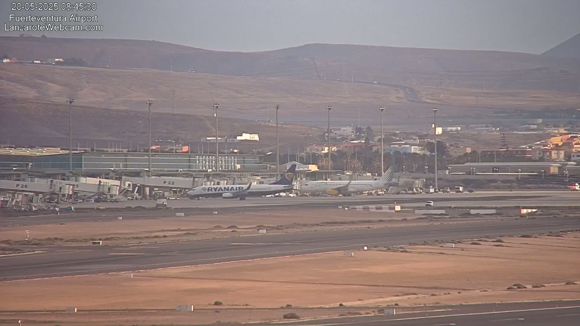 An airport tarmac with two airplanes, one near a jet bridge, under hazy, overcast skies with a hilly, desert-like landscape in the background.