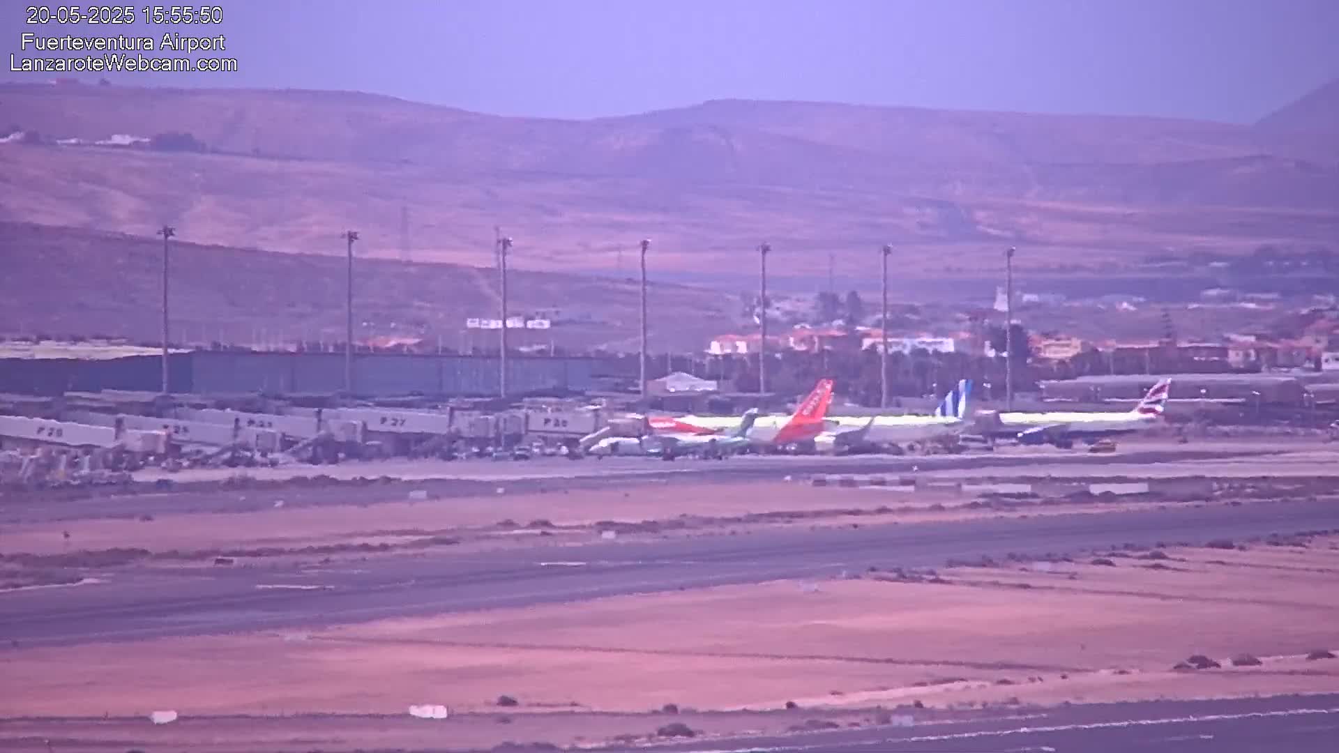Several airplanes are parked at an airport tarmac under a hazy sky, with distant brown hills visible in the background.