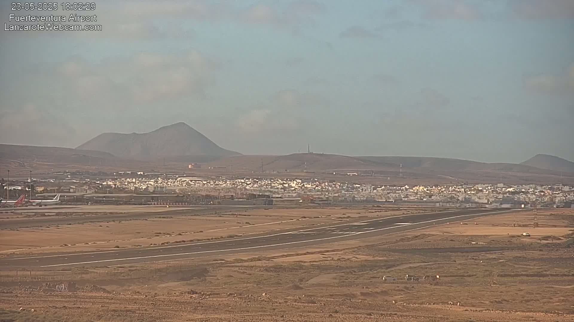 A long runway at an airport stretches across a flat, arid landscape under a hazy sky, with a small town and distant volcanic mountains visible in the background.