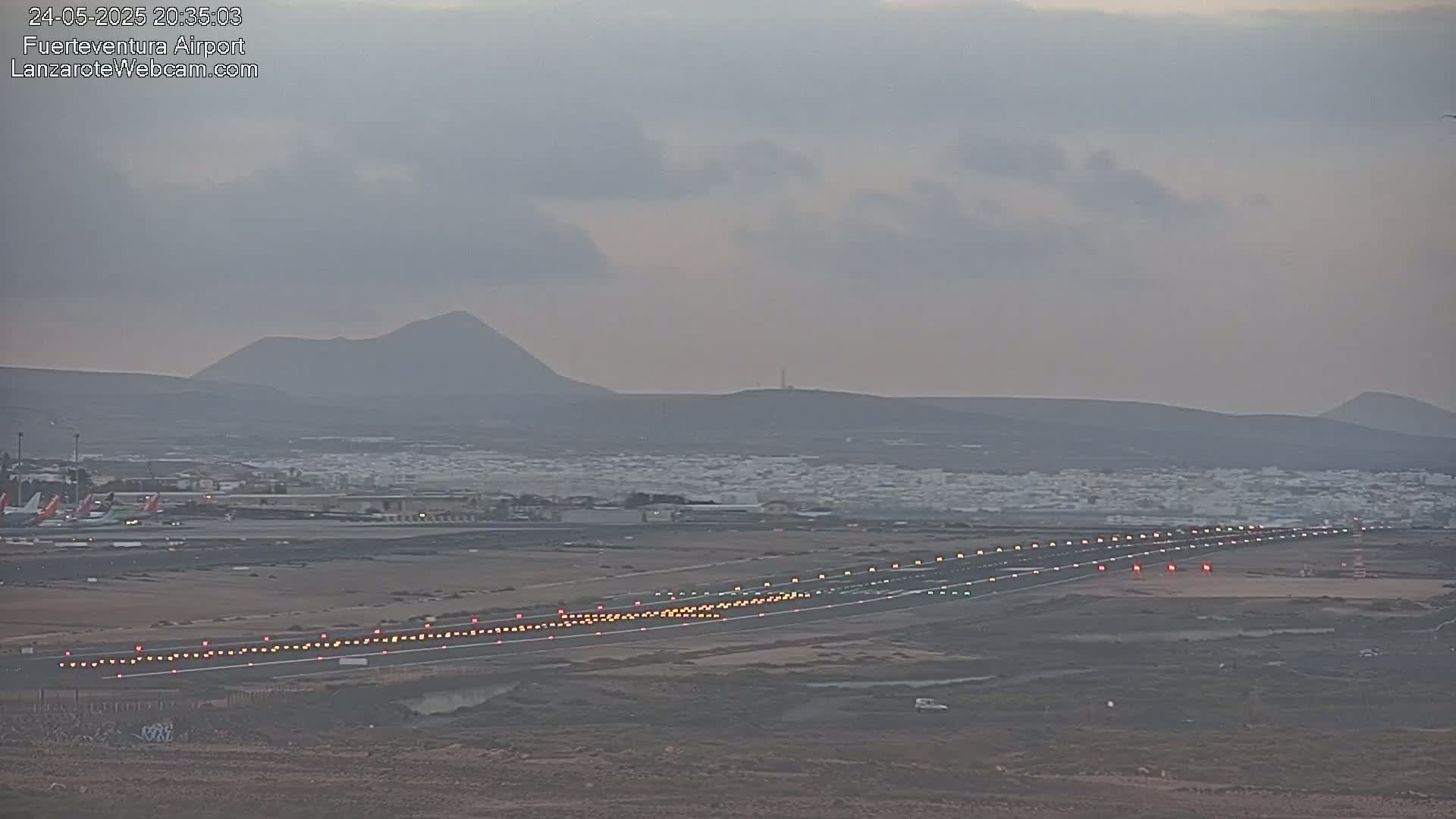 An airport runway with lights illuminated, airplanes parked on the tarmac, and distant volcanic hills under an overcast sky.
