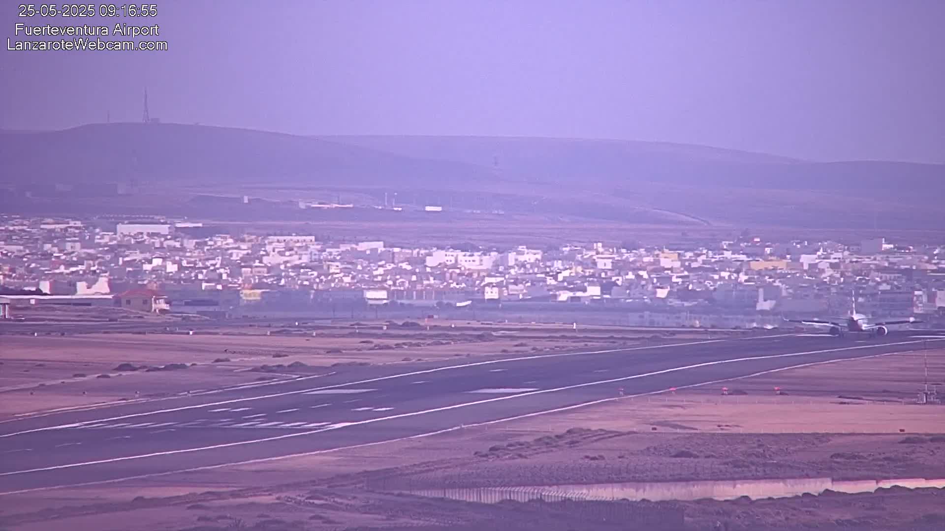 An airplane taxis on a runway toward a distant town under a hazy, purplish sky.