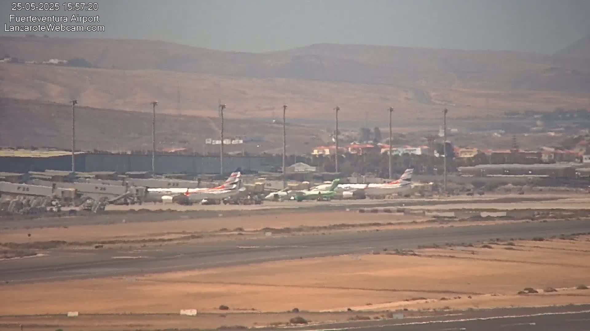 Several airplanes are parked on an airport tarmac under hazy conditions, with a dry, brown landscape and distant hills visible in the background.