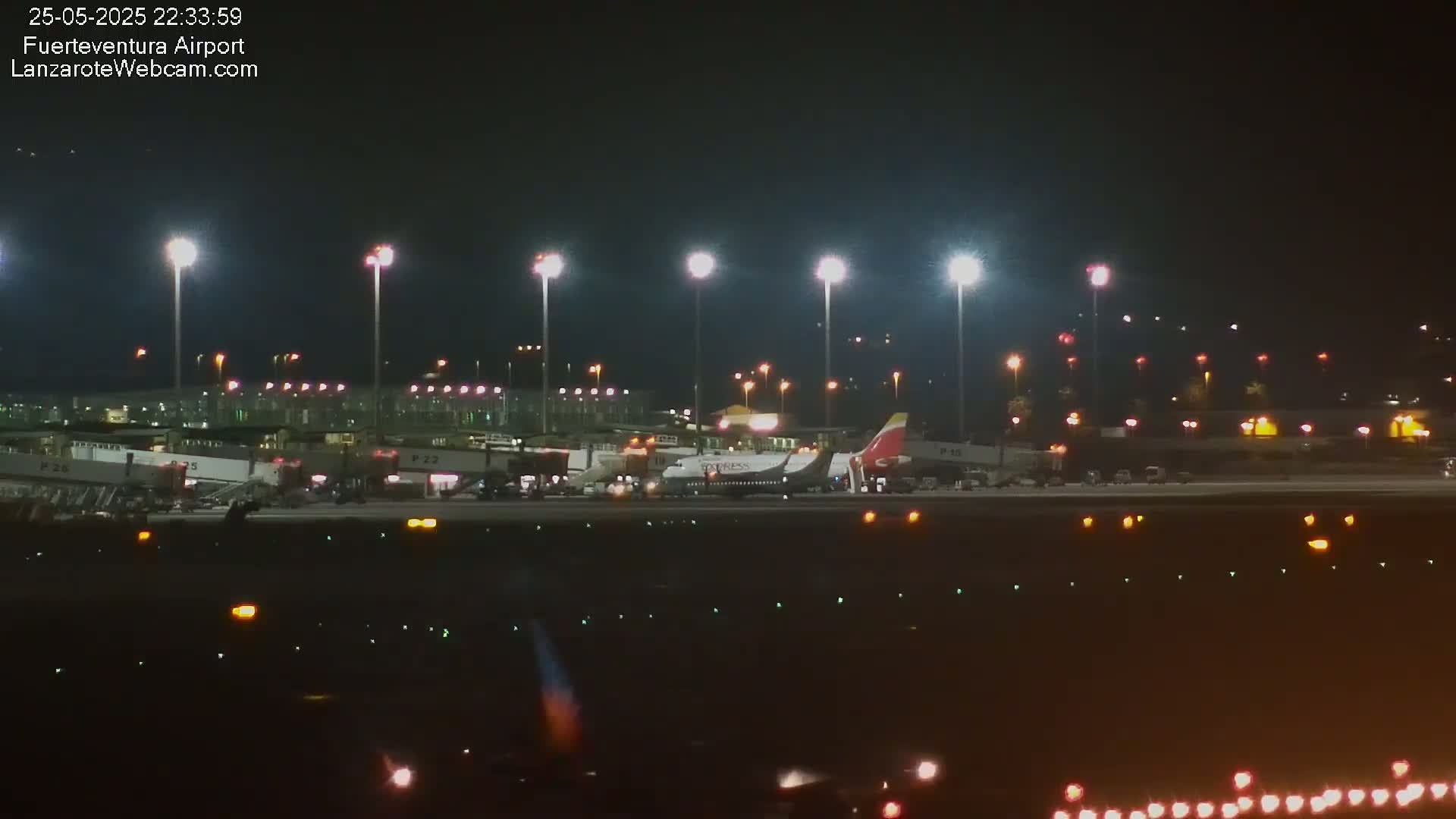 A brightly lit airport tarmac at night shows several airplanes parked near gates, with ground support equipment visible.