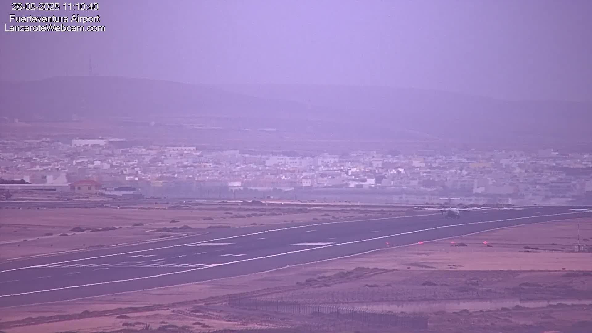 A hazy view shows an airplane taxiing on a runway towards a town in the distance.