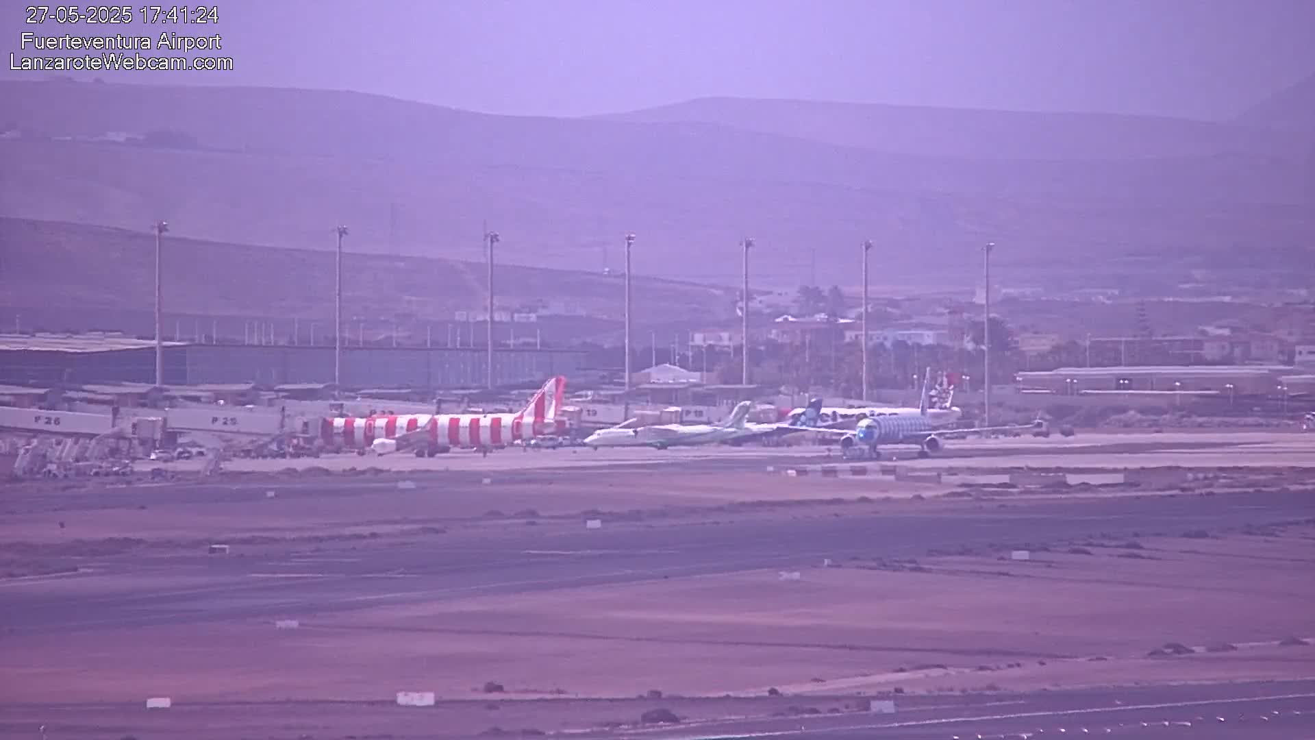 Several airplanes are parked on an airport tarmac under hazy conditions with mountains in the background.