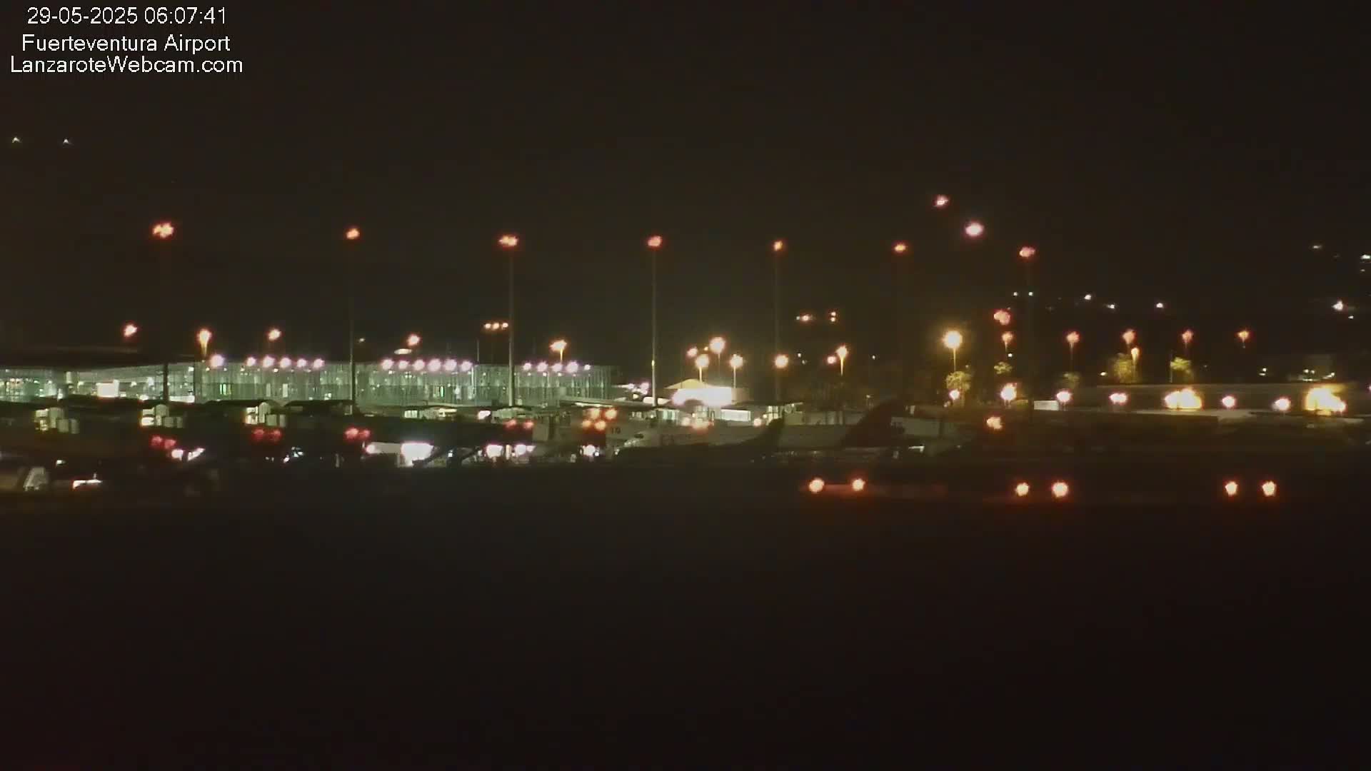 A nighttime view of an airport terminal and tarmac with numerous lights illuminating planes and ground vehicles.