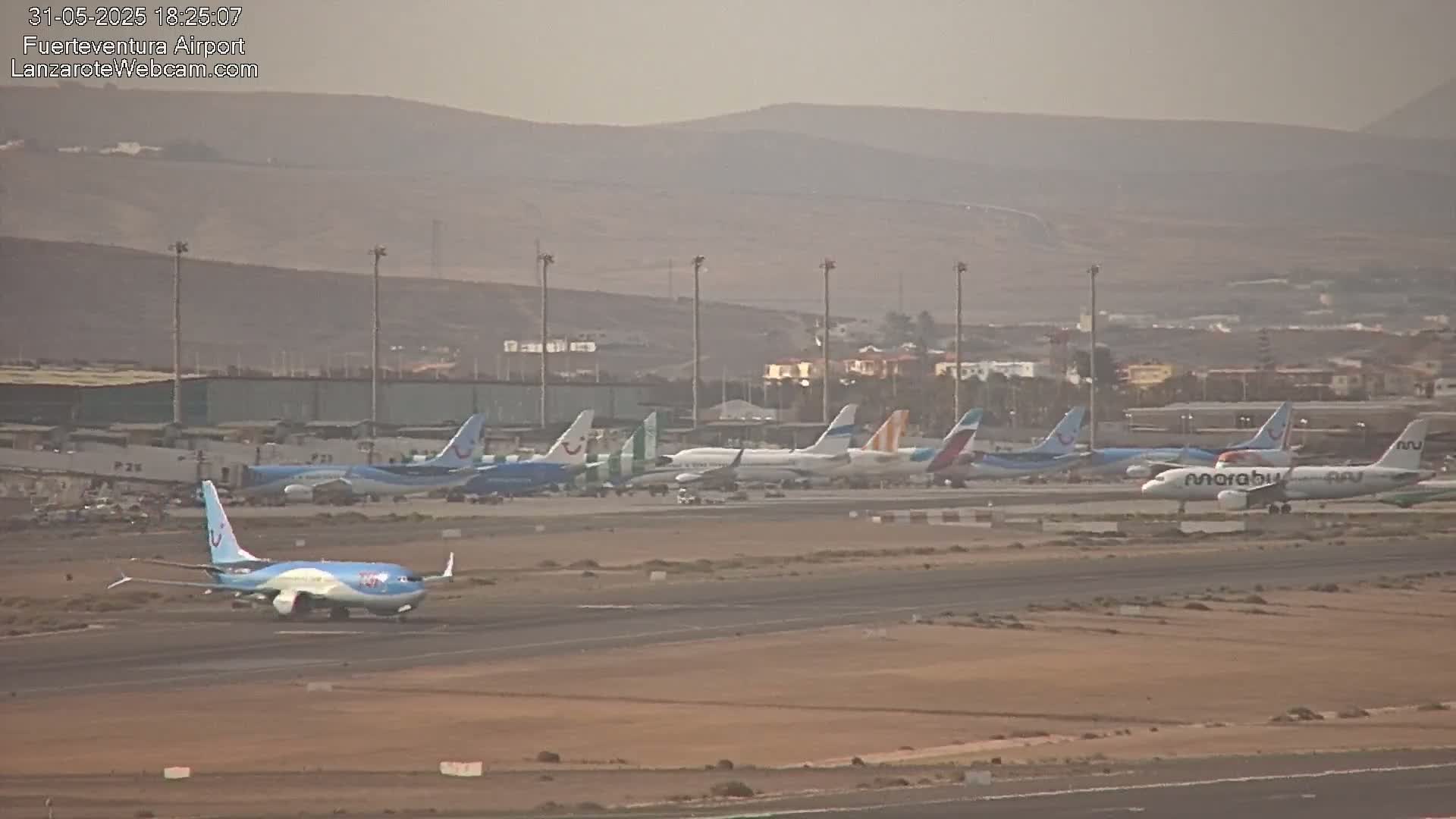 Multiple airplanes are parked on a tarmac at an airport under hazy conditions, with a mountainous landscape visible in the background.