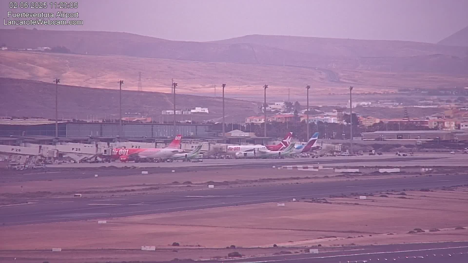 Several airplanes are parked at an airport tarmac under an overcast sky, with hills visible in the background.