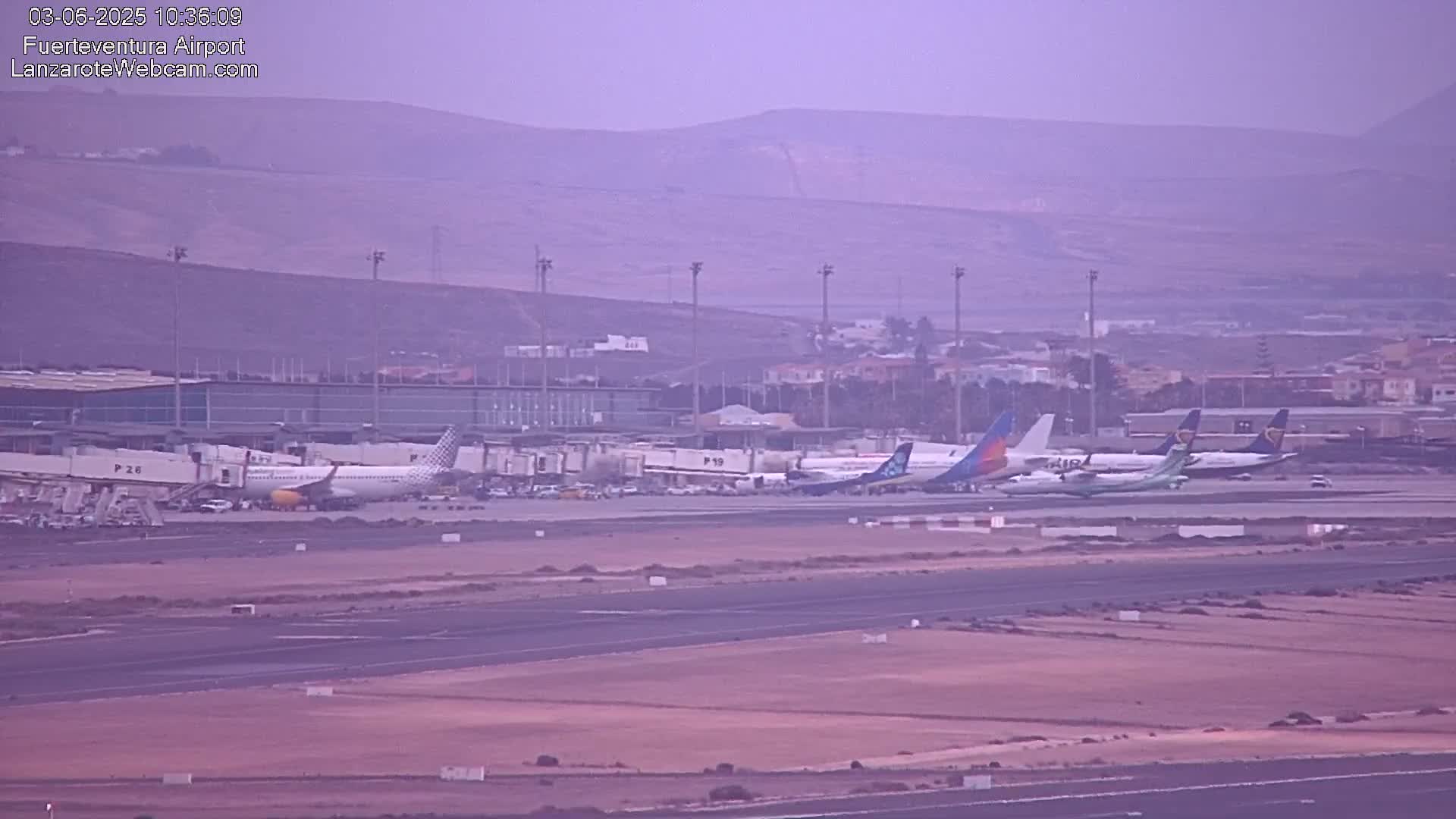 Several airplanes are parked at an airport on a hazy day, with low-lying hills visible in the distance.