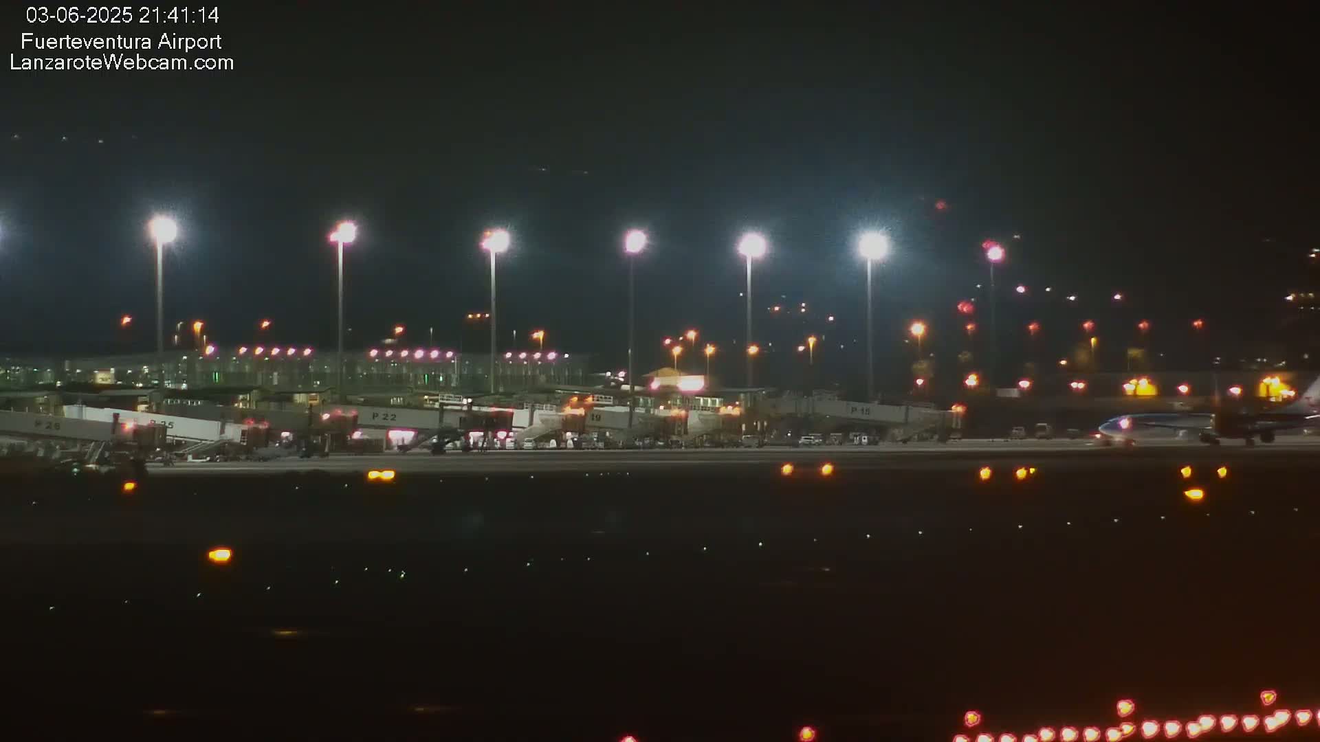 A nighttime view of an airport tarmac with several planes at gates, illuminated by bright lights.
