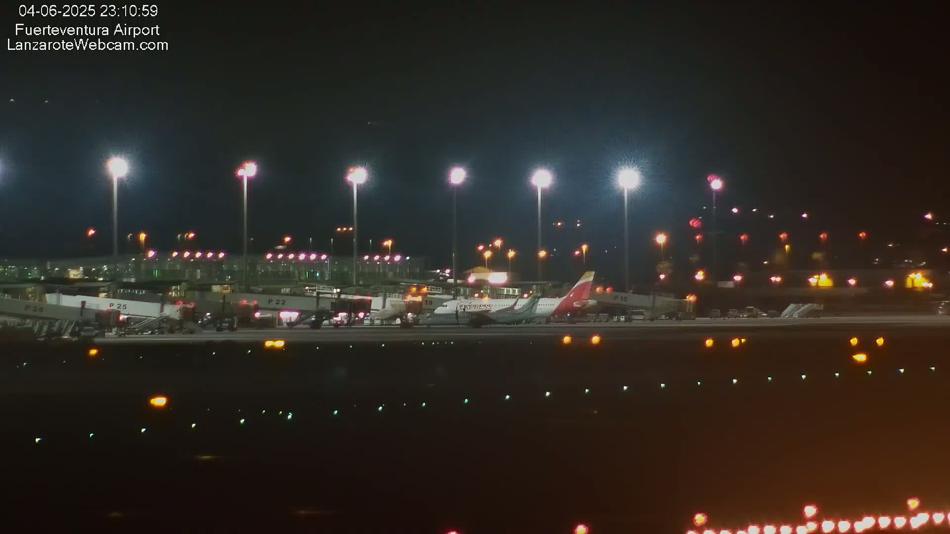 A nighttime view of an airport tarmac with several airplanes and jet bridges illuminated by bright lights.