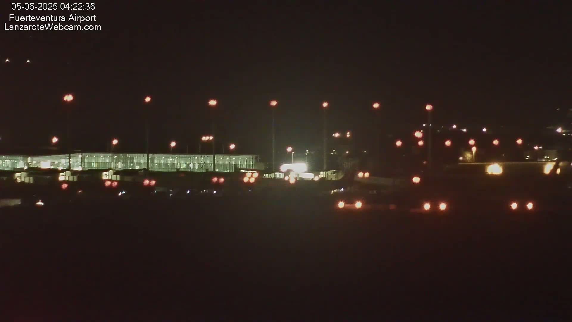 A brightly lit airport terminal at night is visible from a distance, with numerous lights illuminating the tarmac and surrounding area.