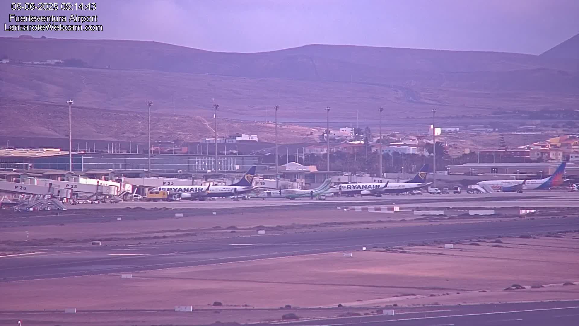 Several airplanes are parked at an airport tarmac under an overcast sky, with a hilly landscape visible in the background.