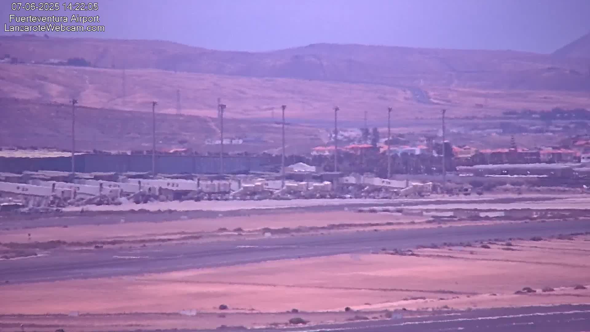 A hazy, long shot of an airport tarmac with ground support equipment and buildings in the background under a hazy sky.