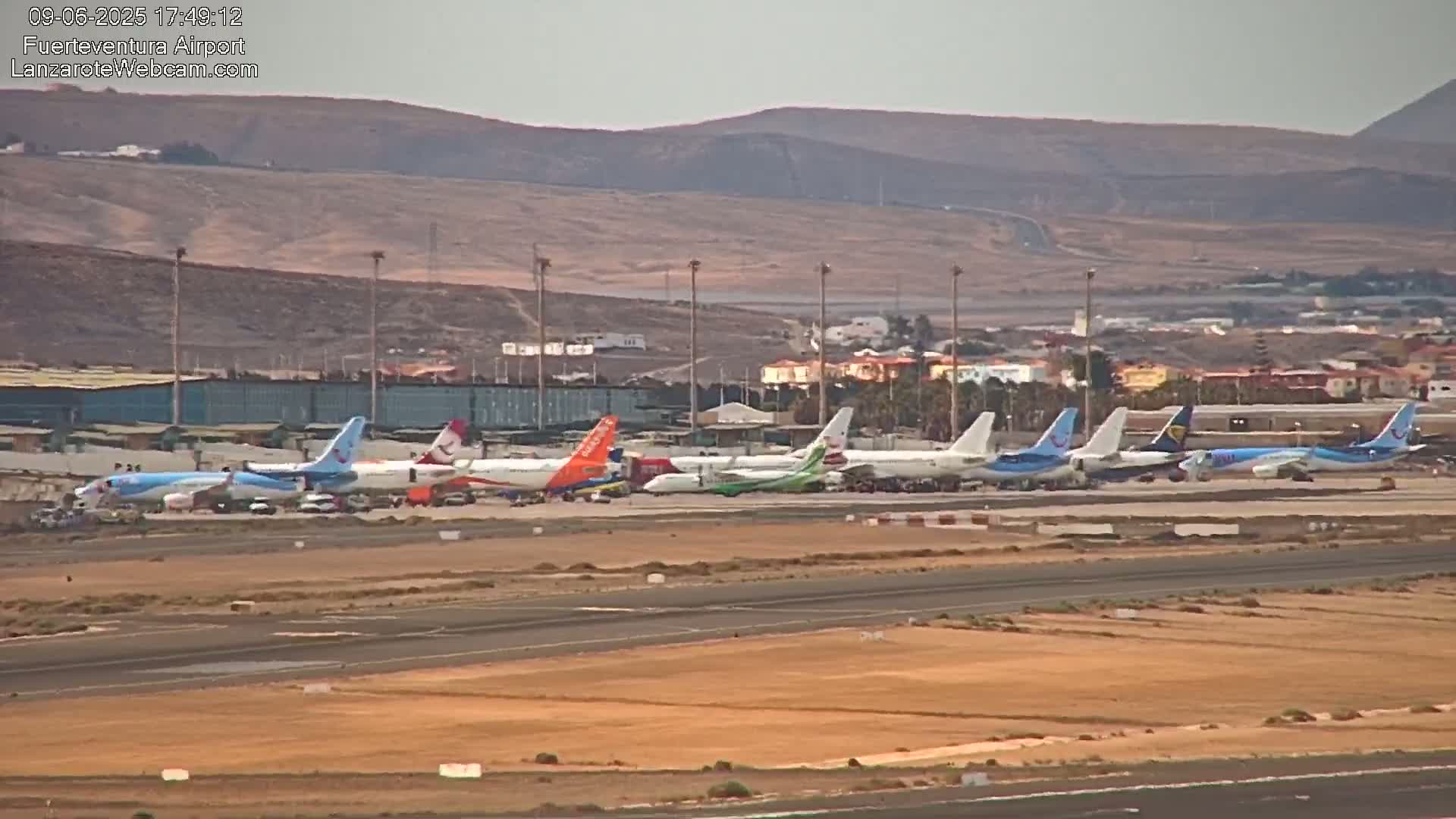 A row of airplanes sits on a tarmac at an airport under a hazy sky, with brown hills visible in the background.