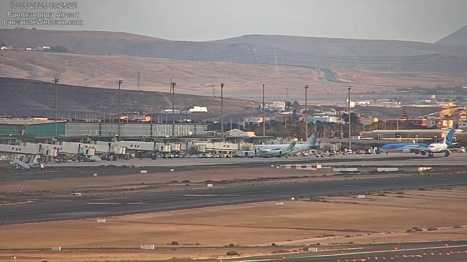 An airport tarmac with several airplanes, including one at a gate and another on the runway, under hazy, sunny conditions with arid, mountainous terrain in the background.