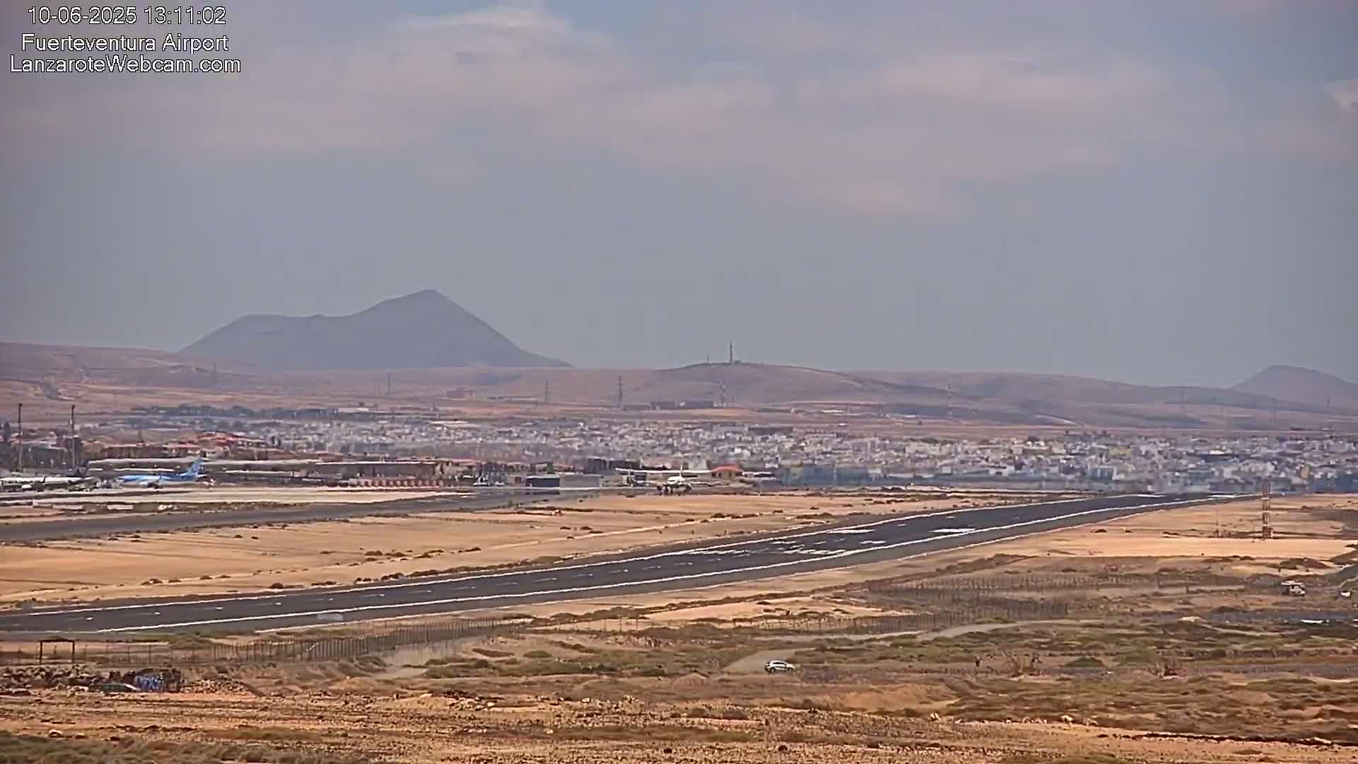 A long airport runway stretches across a flat, arid landscape under a hazy sky, with a small town and distant volcanic hills in the background.