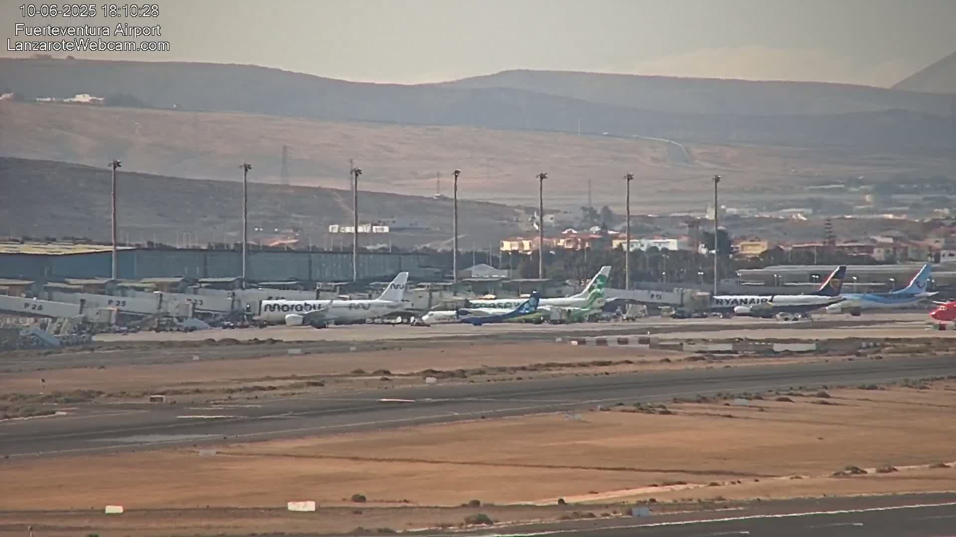 Several airplanes are parked at an airport tarmac on a hazy day with a mountainous background.