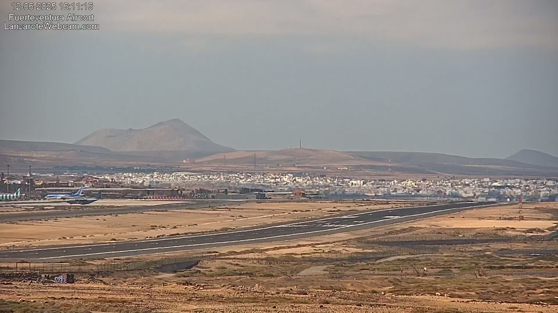 A plane taxis on a runway at an airport situated in a desert-like area with low hills and a town visible in the background under hazy conditions.