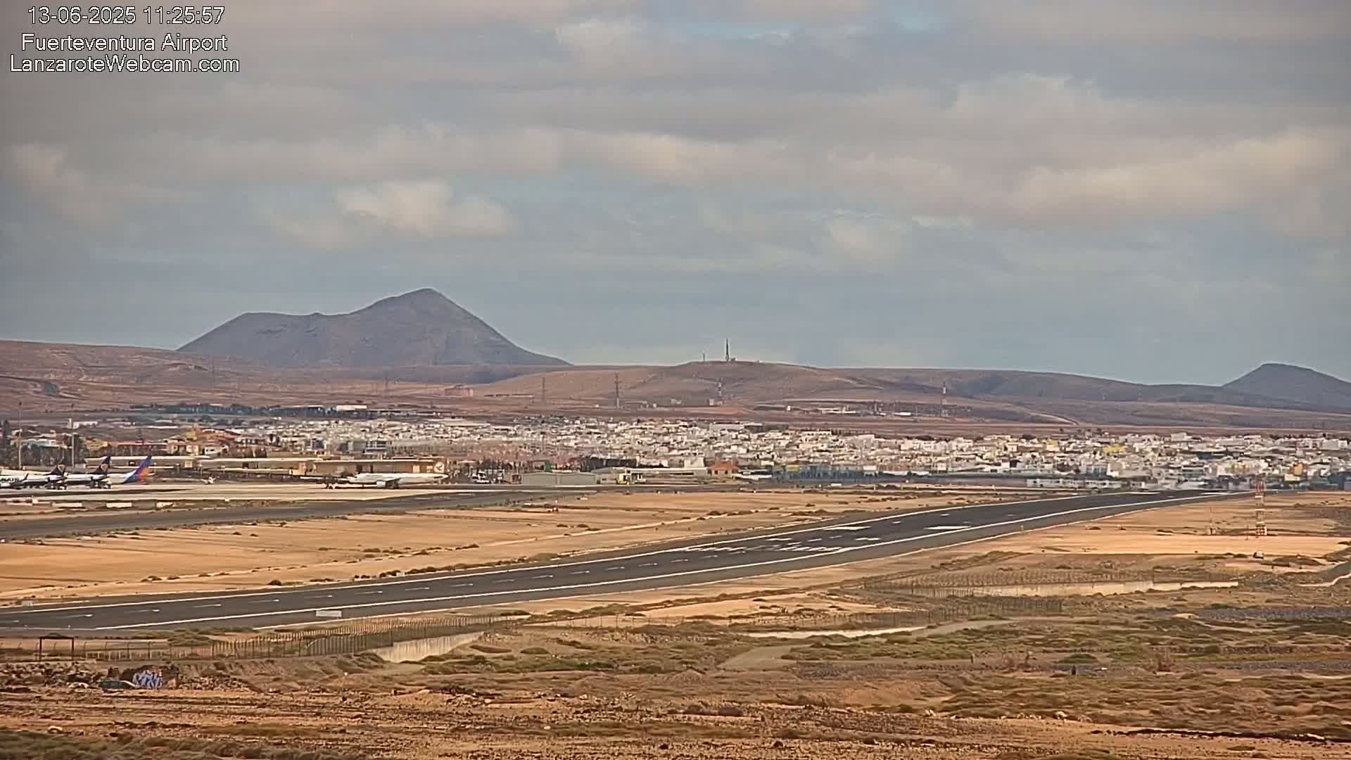 An airport runway with several planes, a small town, and volcanic hills in the background under a mostly cloudy sky.