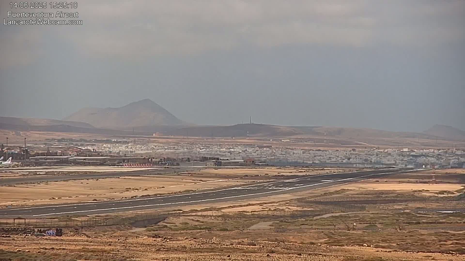 A long airport runway stretches across a flat, dry landscape under a hazy sky, with distant mountains visible on the horizon.