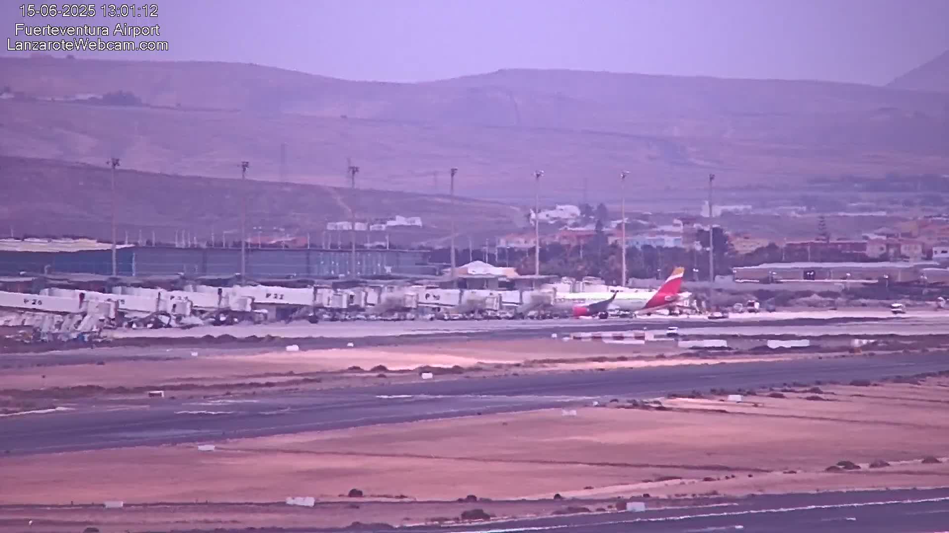 An airplane is parked on an airport tarmac under hazy conditions, with several ground support vehicles nearby and a distant view of hills.
