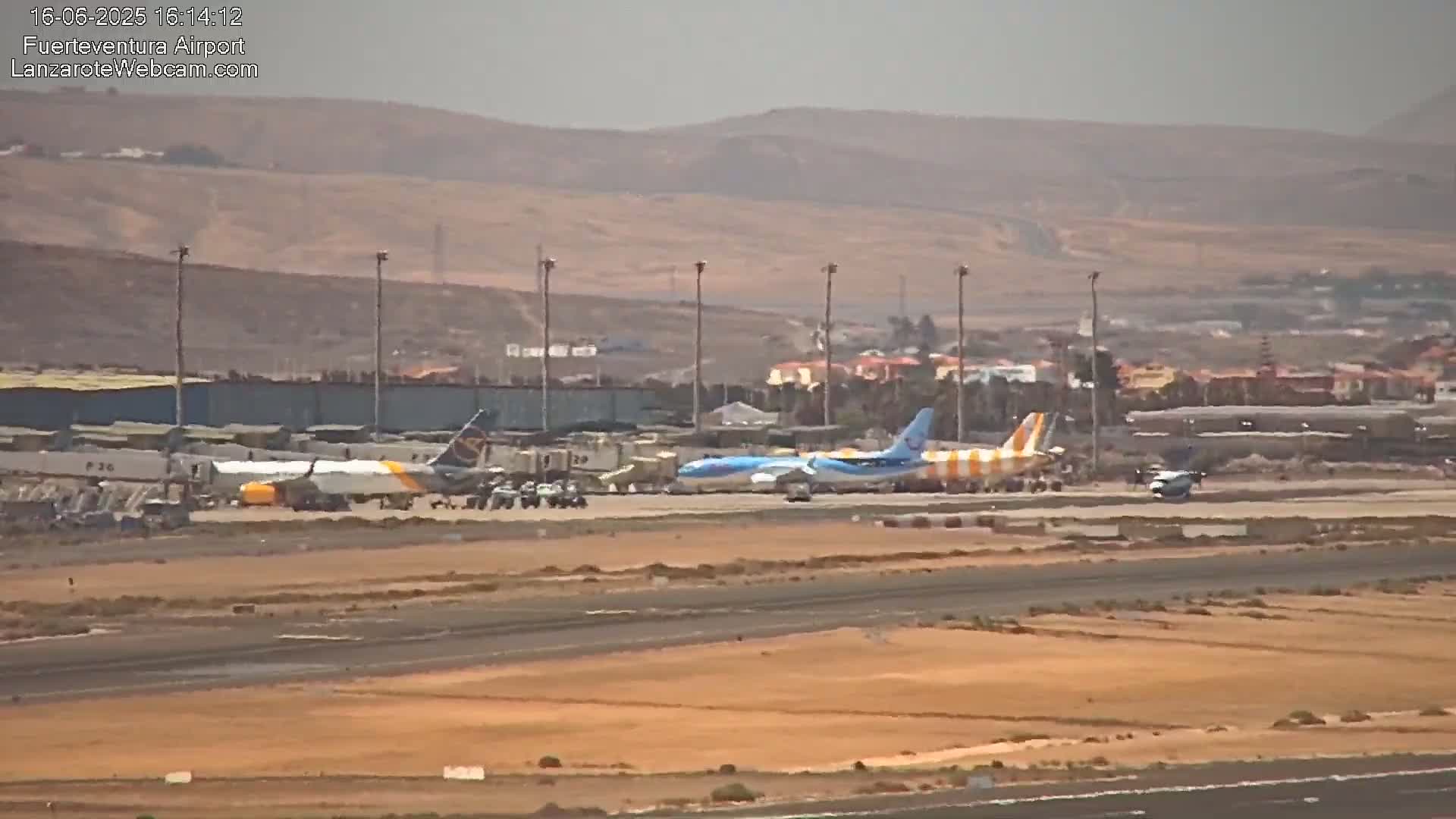 Several airplanes are parked on an airport tarmac under hazy conditions, with a mountainous, dry landscape visible in the background.