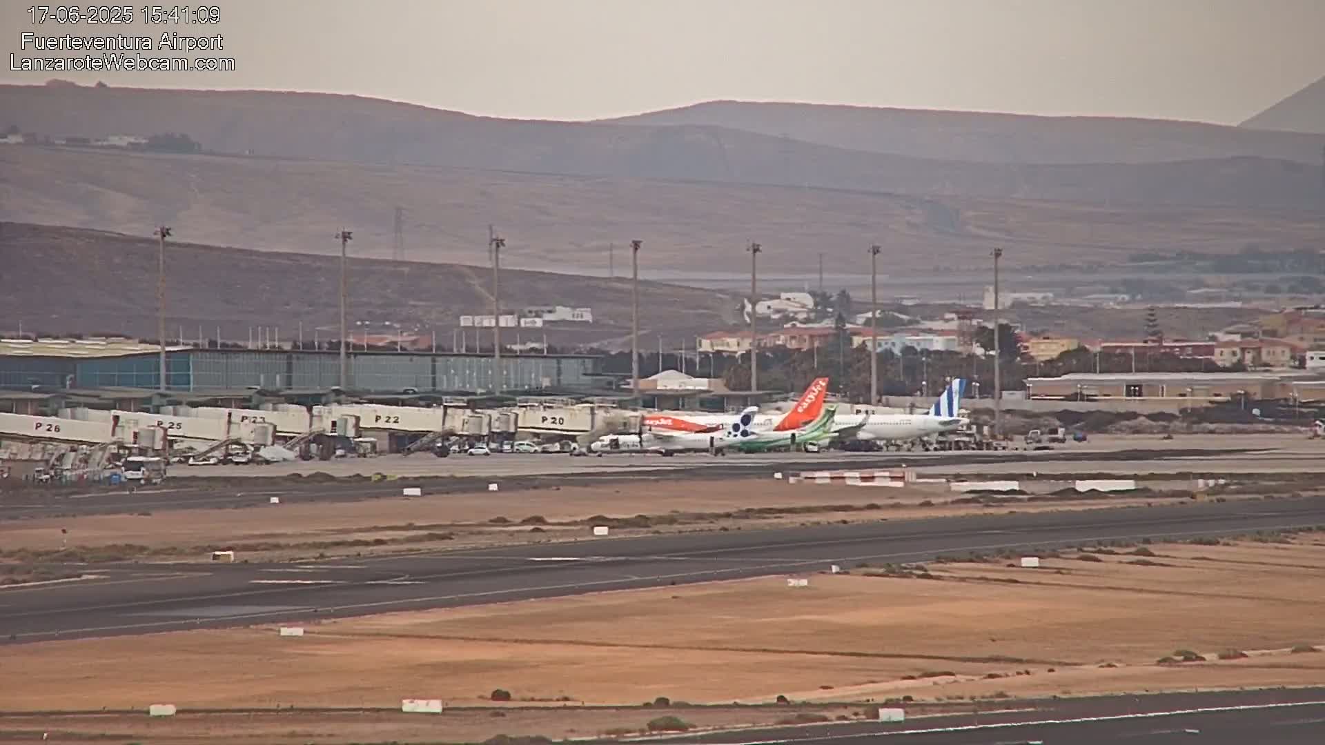 Several airplanes are parked at an airport terminal under an overcast sky, with arid hills visible in the background.