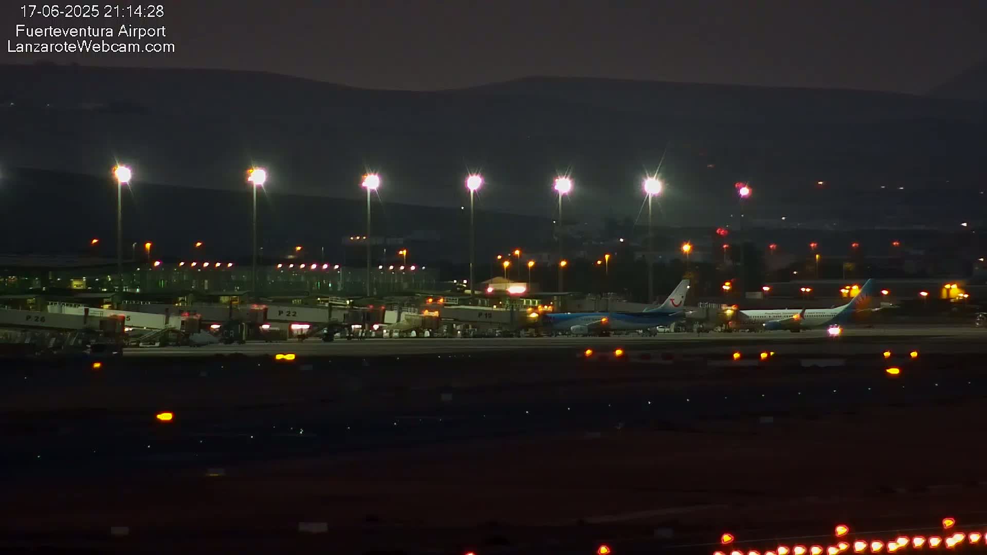 At night, two airplanes are parked at an airport terminal under bright lights, with hills visible in the background.