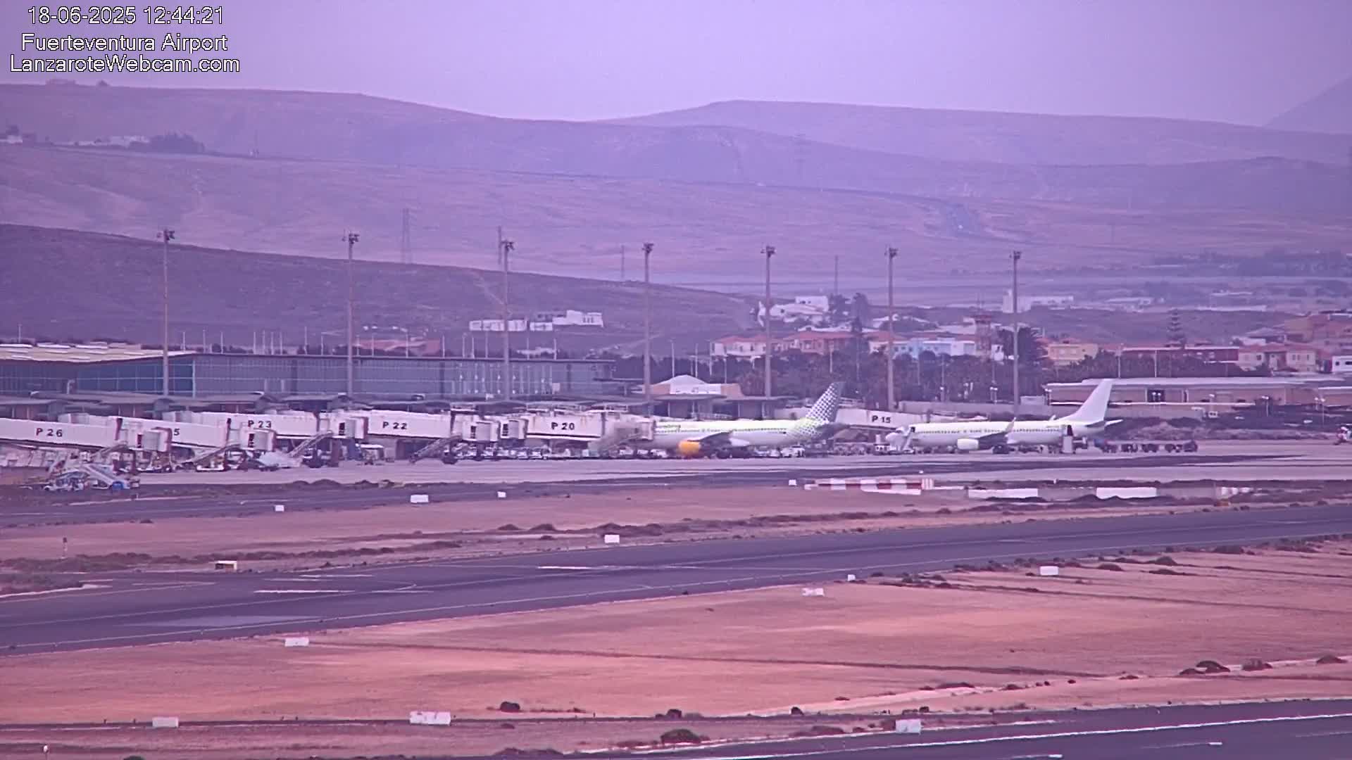 A passenger airplane is parked at an airport terminal under hazy conditions, with mountains visible in the background.
