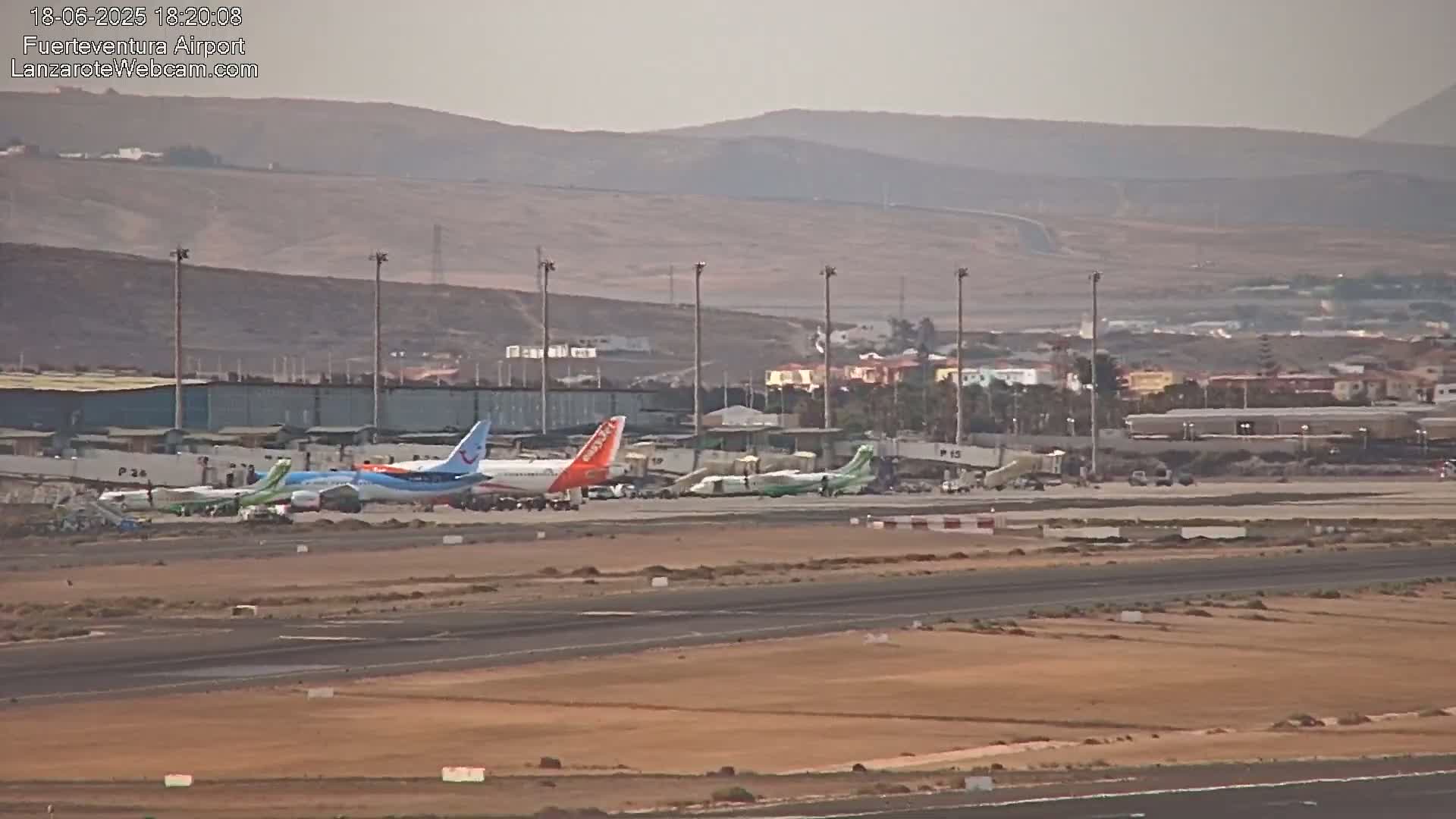 Several airplanes are parked at an airport tarmac on a hazy day, with a distant view of low-lying hills and buildings.