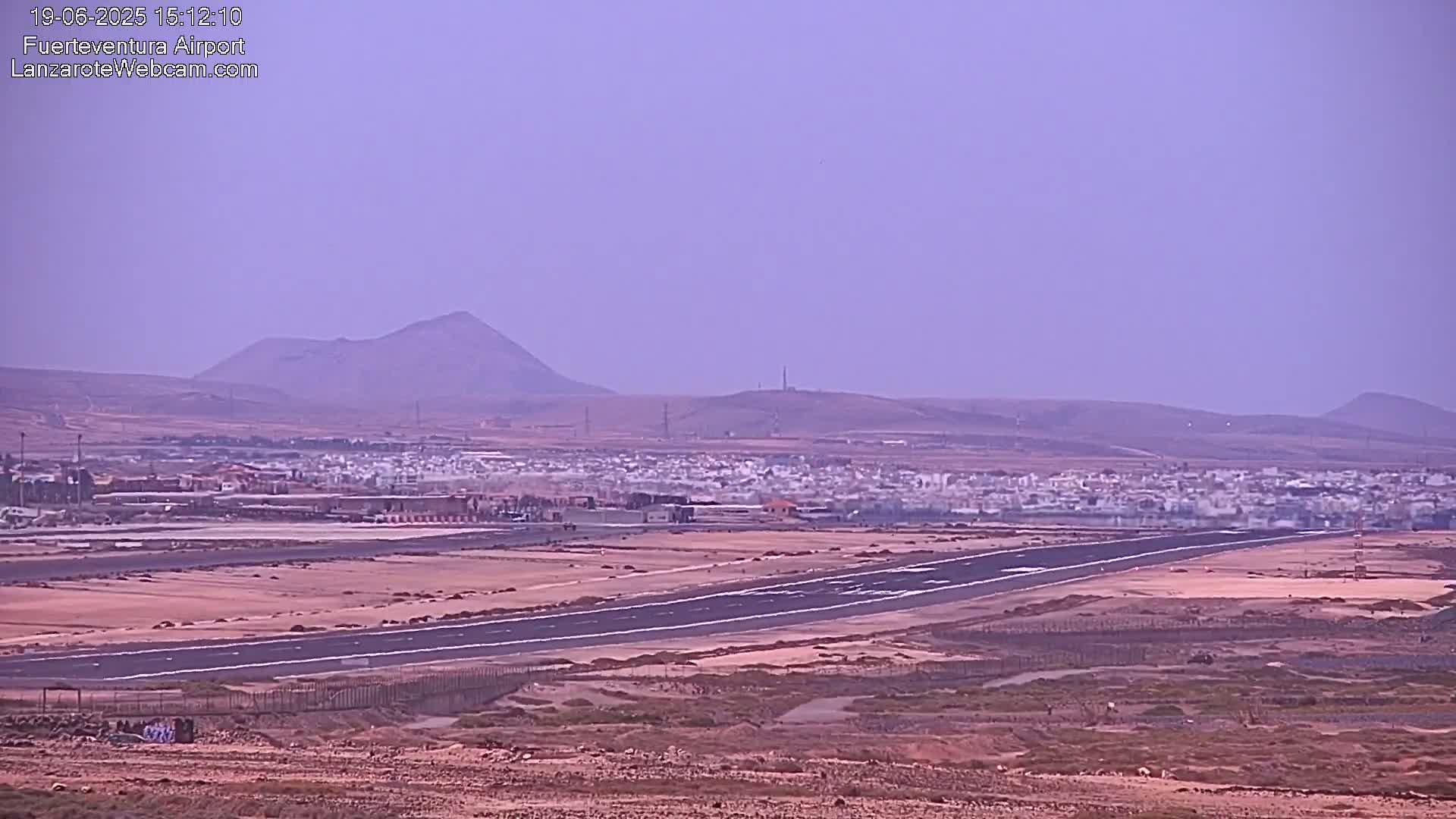 A long airport runway stretches across a flat, arid landscape under a hazy, light purple sky, with distant hills and a small town visible in the background.