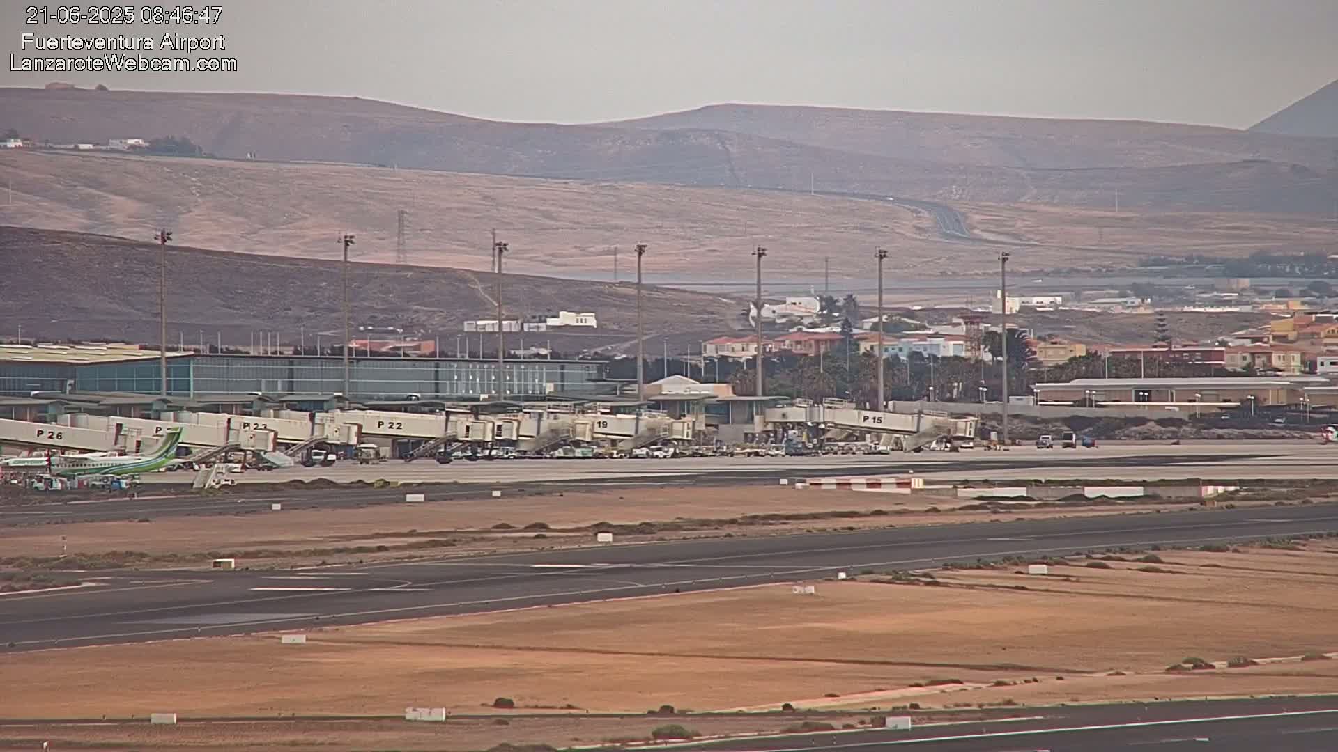 An airport tarmac with several airplanes at gates, surrounded by buildings and brown, hilly terrain under hazy skies.